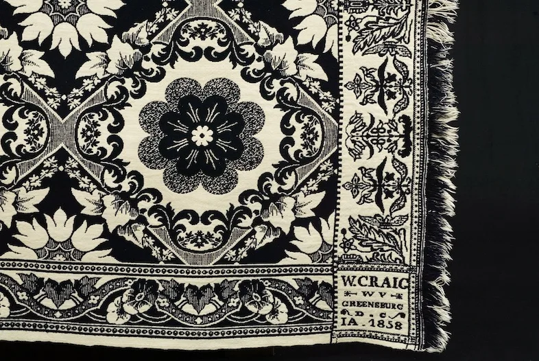 Corner of a dark indigo and white jacquard coverlet showing a large flower medallion in a foliate lozenge, a scrolling side border, a knotted fringe along the edge and a small lettered block reading W. Craig, Greensburg, and the date 1858