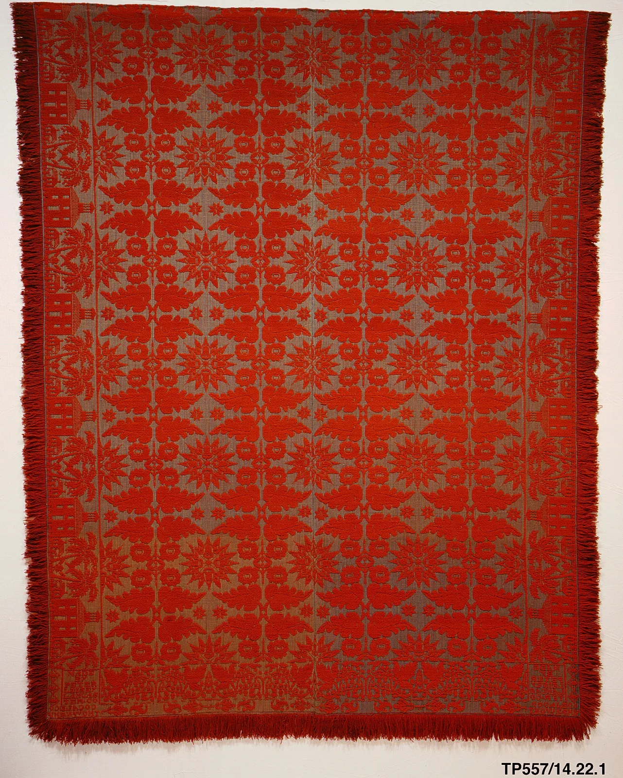 Coverlet in strong orange-red and a muted olive-grey, patterned all over with rows of small floral medallions and stars in a regular grid