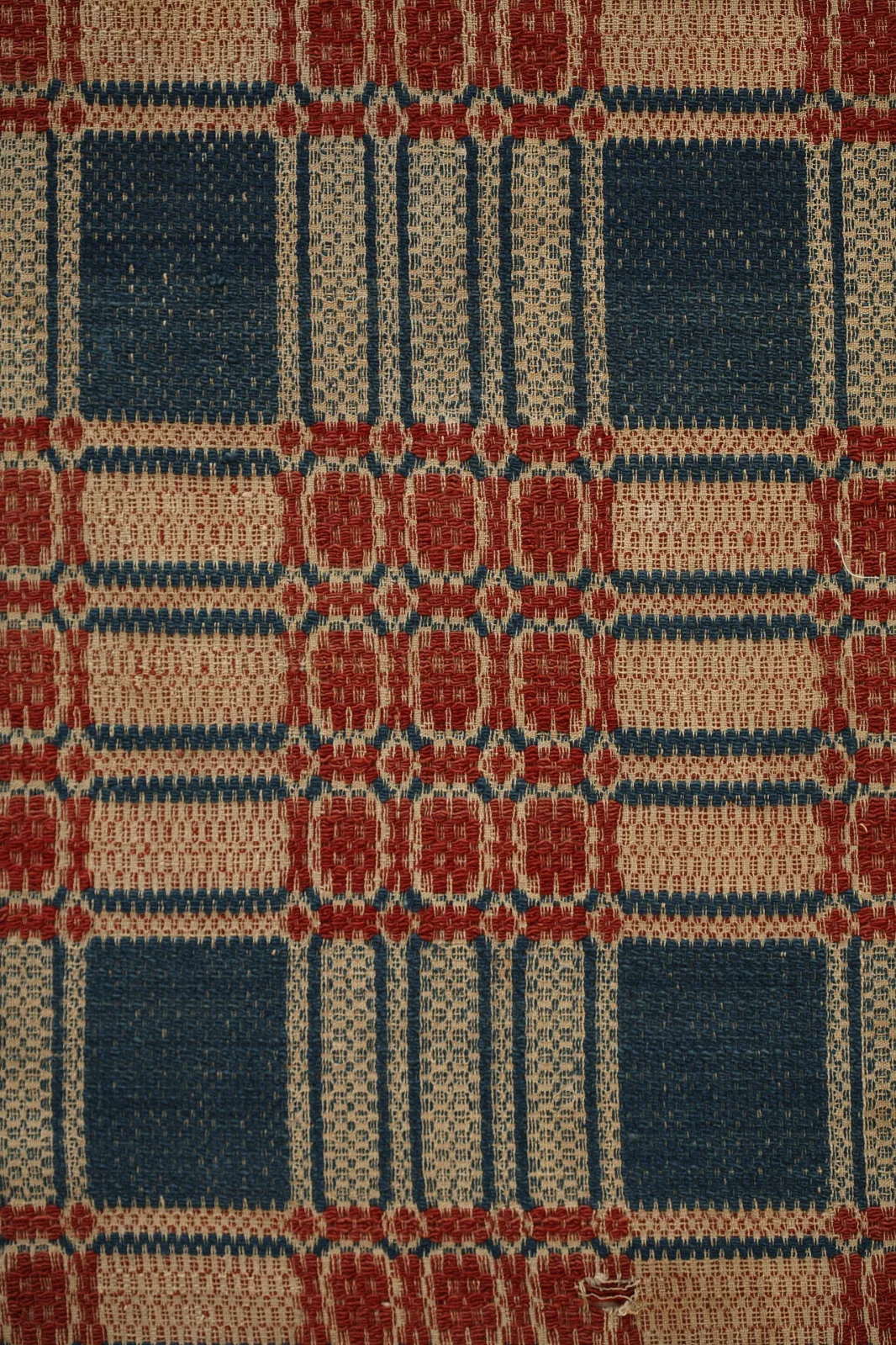 Close view of a worn overshot coverlet in dark rose, medium blue and beige, showing blocks of coloured wool floating loosely across the surface over a finer plain-woven ground visible between them