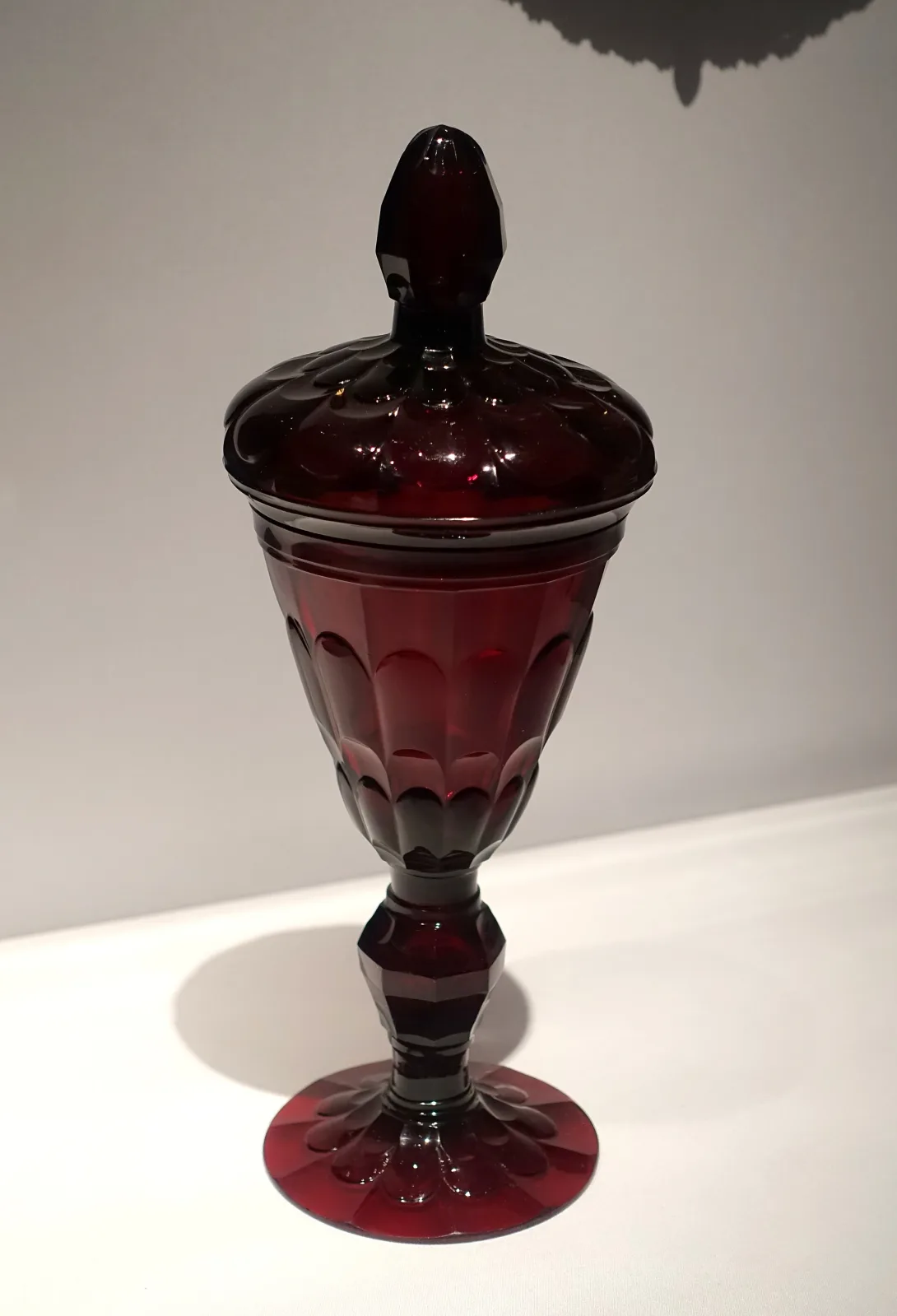 Tall covered goblet in very dark red glass, cut with flat vertical panels on the bowl and a faceted knop on the stem, with a pointed faceted finial on the lid