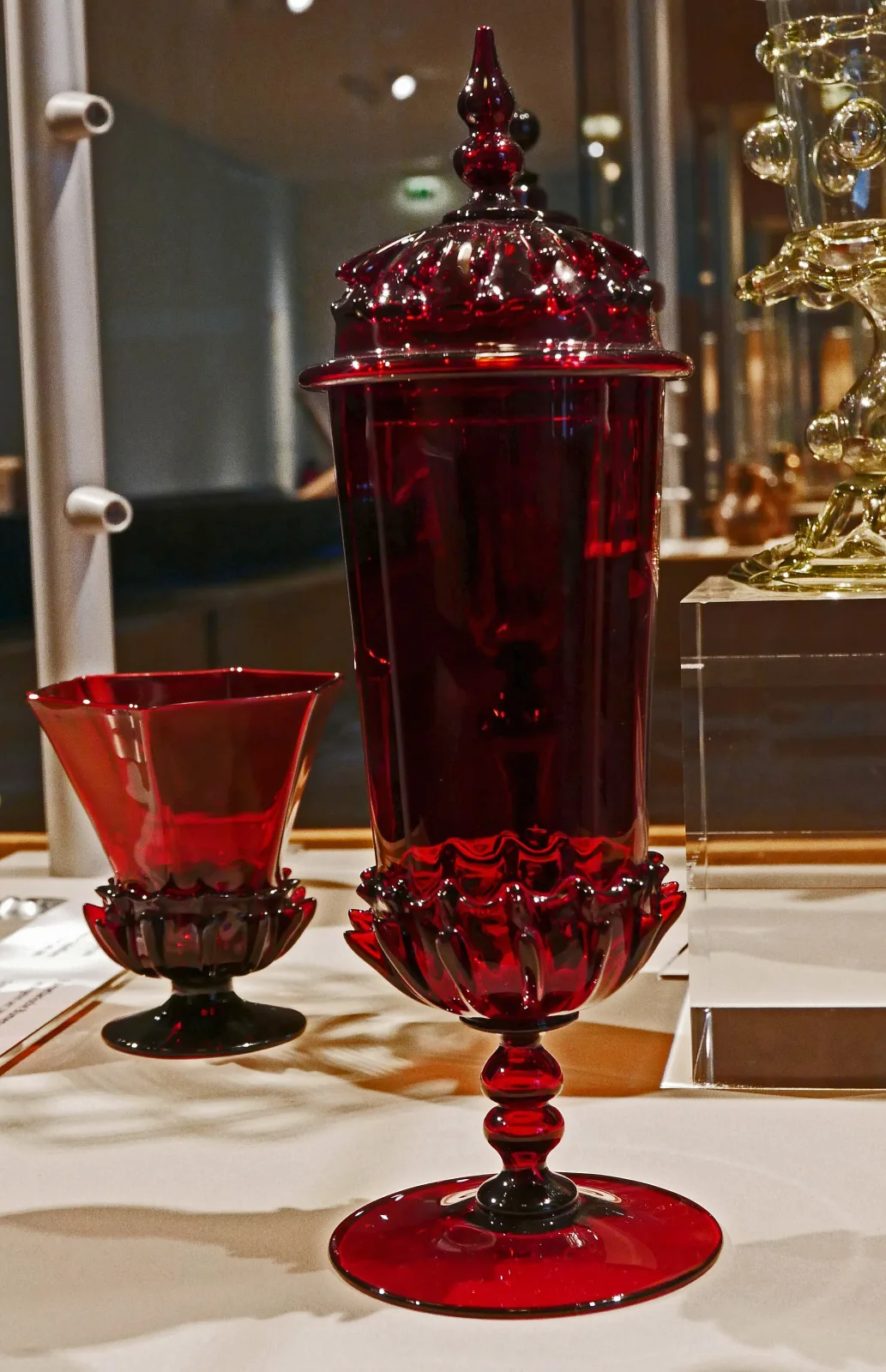 Tall covered ruby glass pokal with a gadrooned and drop-applied collar at the base of the bowl, a knopped stem on a wide plain foot and a spired finial, beside a smaller matching beaker in a museum case