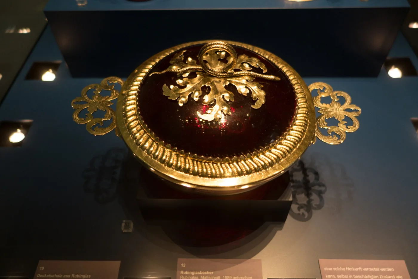 Covered bowl of very dark red glass seen from above, banded and topped with elaborate pierced and chased gold mounts with foliate handles