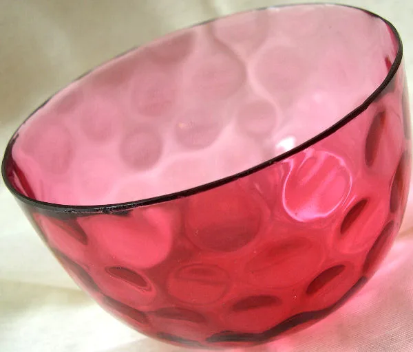 Deep rose-red glass bowl photographed at an angle, its walls covered in regular rows of oval concave indentations that read distinctly paler than the glass around them