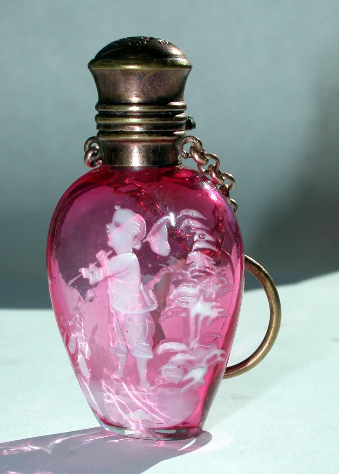 Small rose-red glass scent flacon painted with a white enamel figure of a child among foliage, fitted with a metal screw cap on a threaded collar with a short chain and a suspension ring
