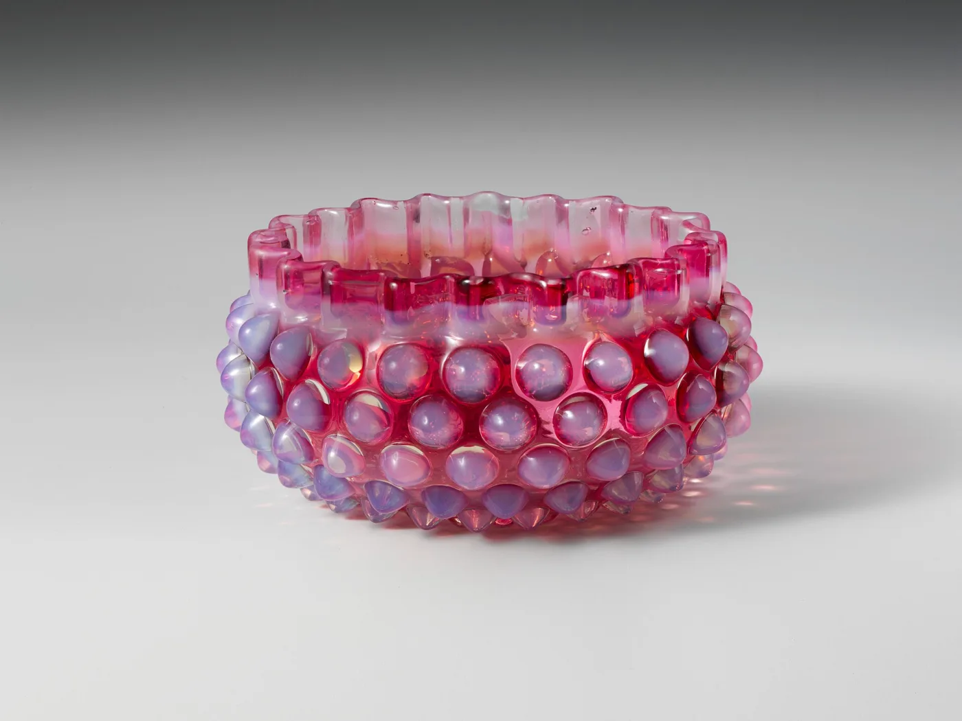 Low rose-red glass bowl covered in rows of raised hobnails whose rounded tips are filled with milky white, below a crimped rim that is also white, with a band of clear deep cranberry between the two