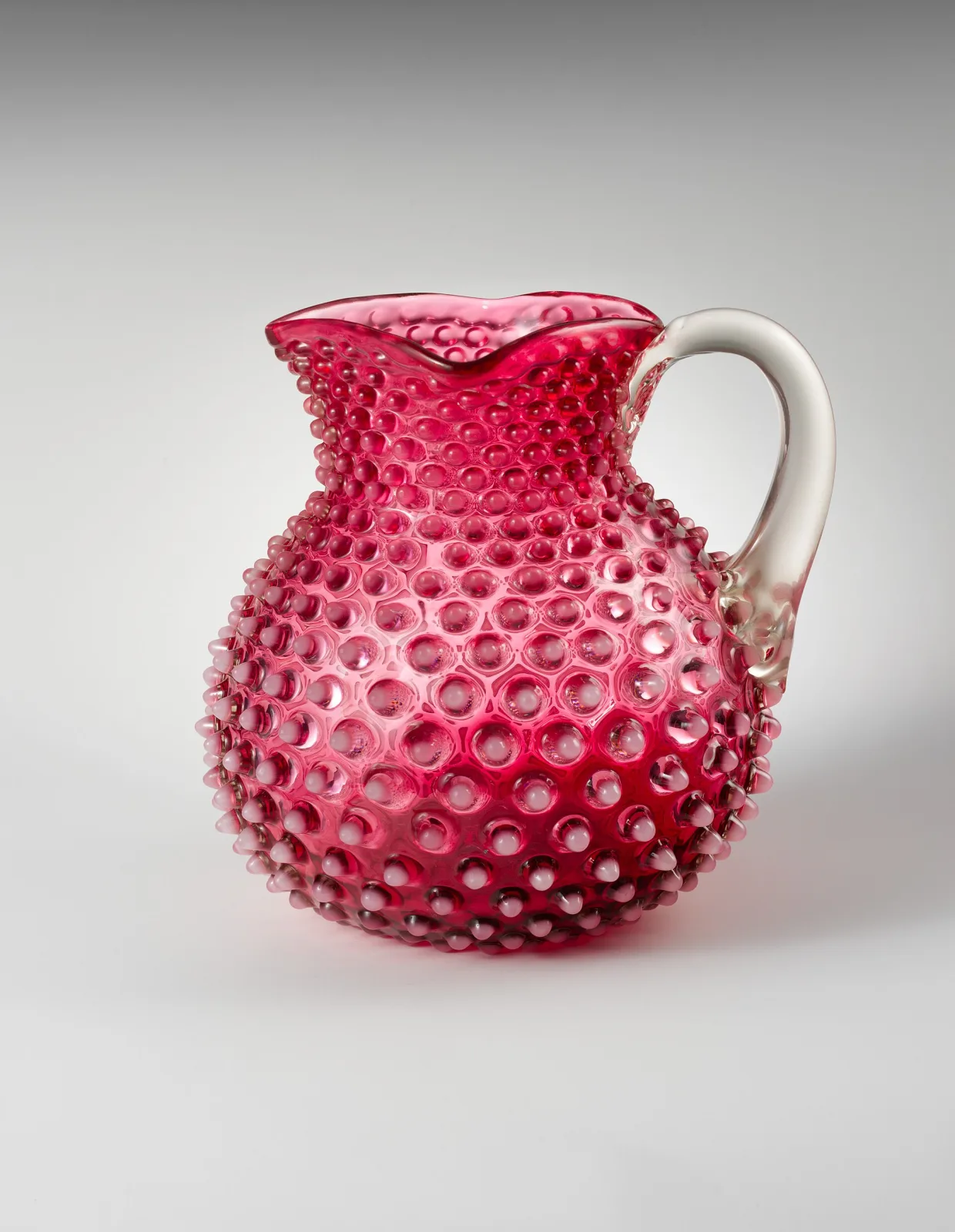 Cranberry glass water pitcher covered in raised hobnails whose tips shade to milky white, with a clear applied glass handle and a crimped rim