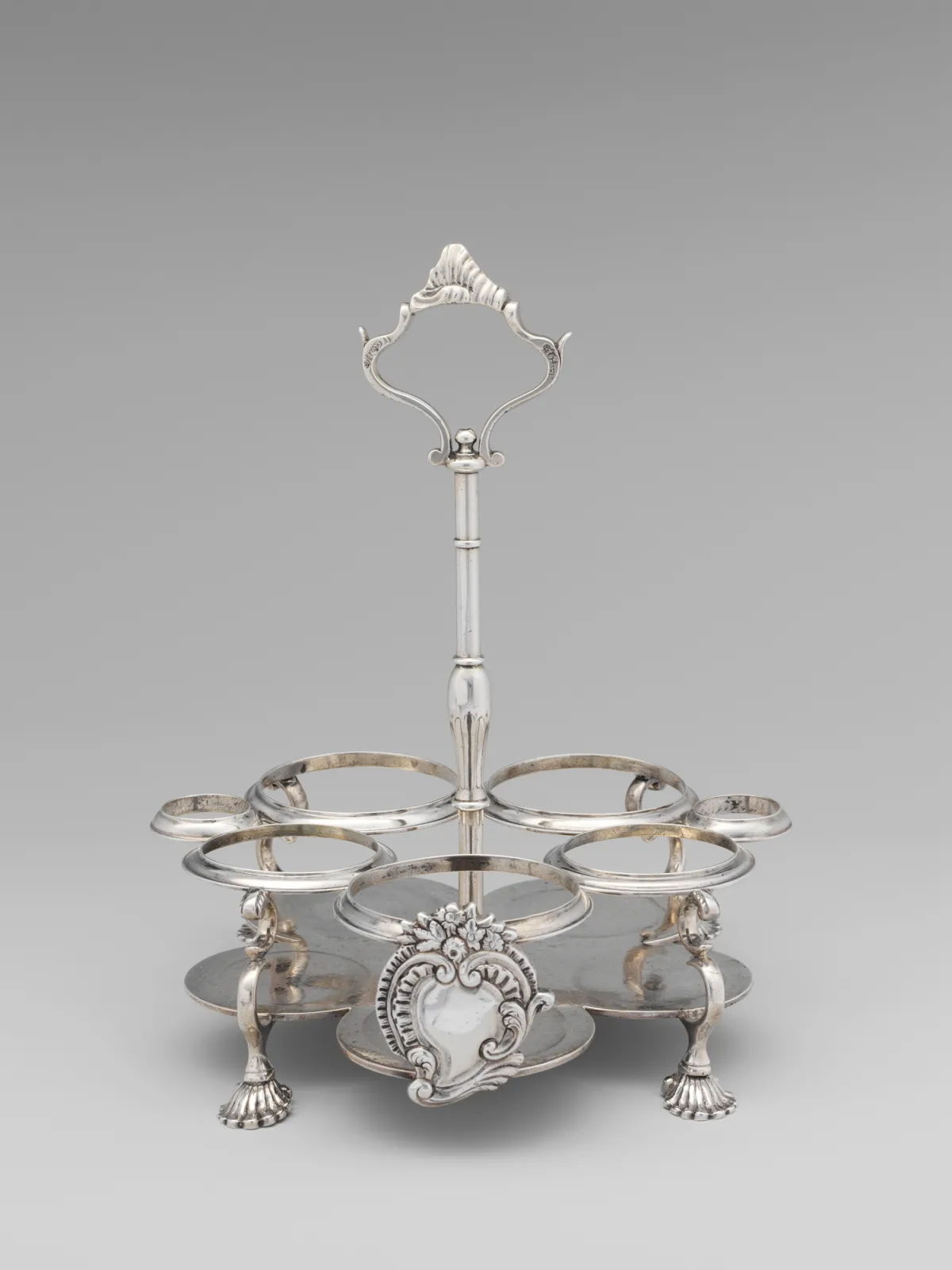 Empty silver cruet frame with seven bottle rings in two sizes — five large and two smaller set lower at the sides — around a central shaped rococo handle on a baluster column, standing on four cast shell feet with a cast cartouche at the front