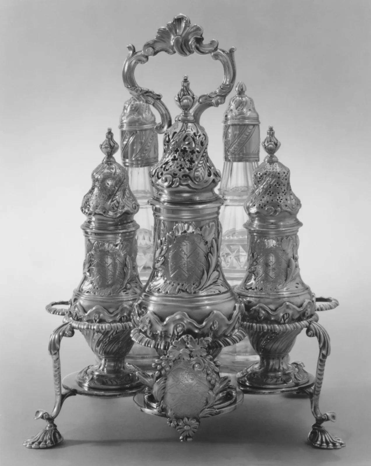 Rococo silver cruet frame on four scrolled feet with a shell-crested handle, holding three pierced-top silver casters engraved with coats of arms and two cut-glass bottles with silver caps