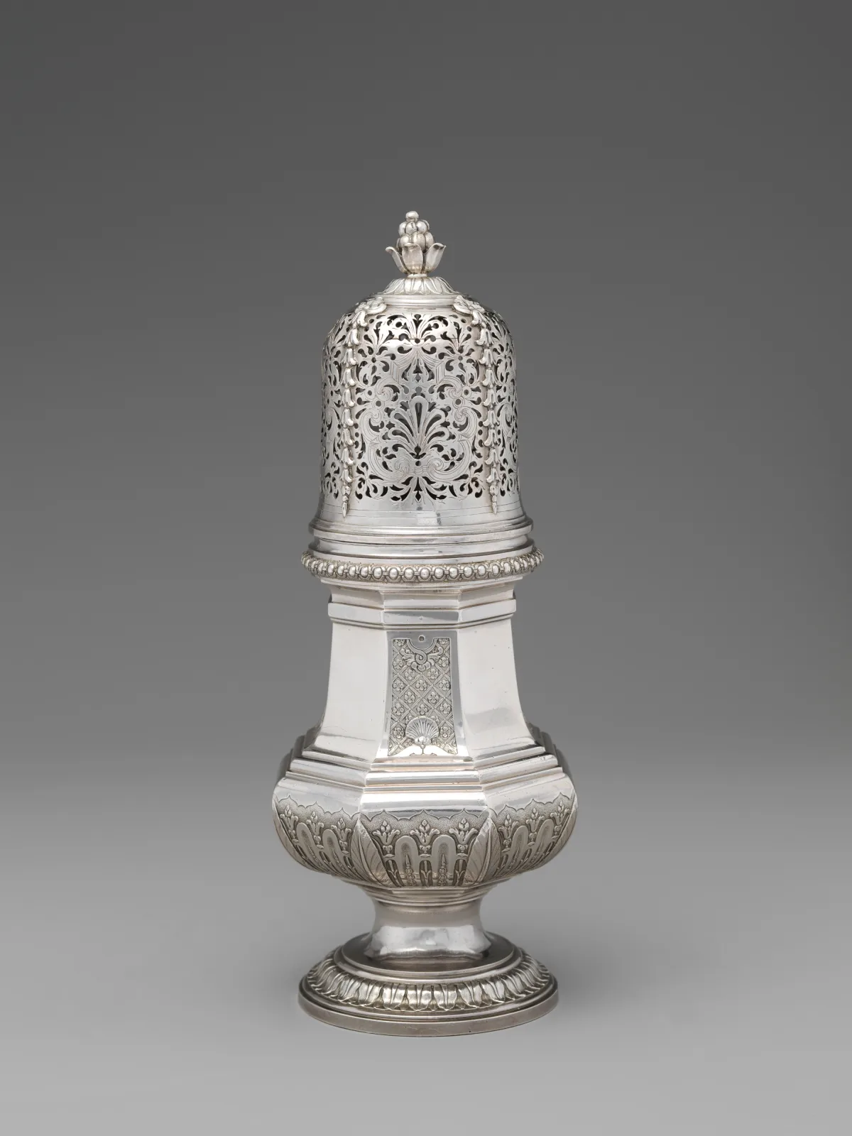 Tall silver sugar caster with a pierced domed cover and a small budded finial above a faceted baluster body decorated with applied strapwork panels, on a stepped moulded foot