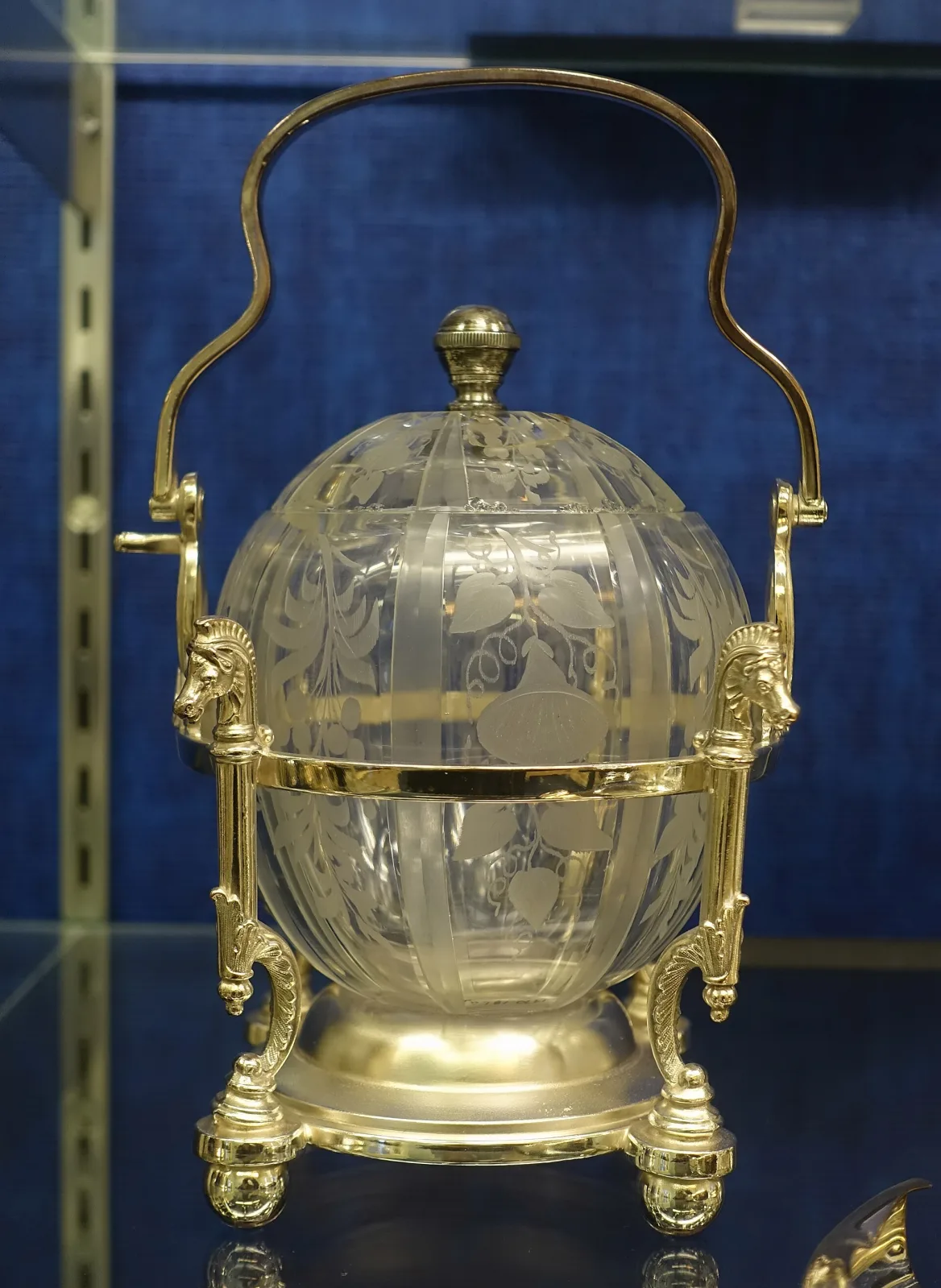 Spherical glass jar engraved with vines and a blank oval reserve, cut in vertical panels and held at its equator by a gilt ring between two supports with animal-head terminals, under a tall swing handle, on a domed gilt foot with four feet