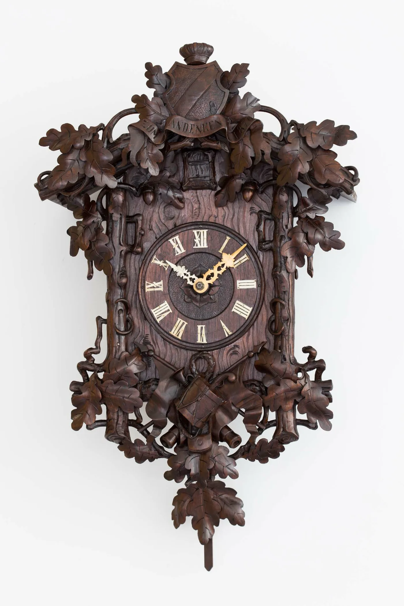 Deeply carved Black Forest cuckoo clock covered in oak leaves and acorns, with a carved crest above the cuckoo door and a drum, helmet and weapons below the dial