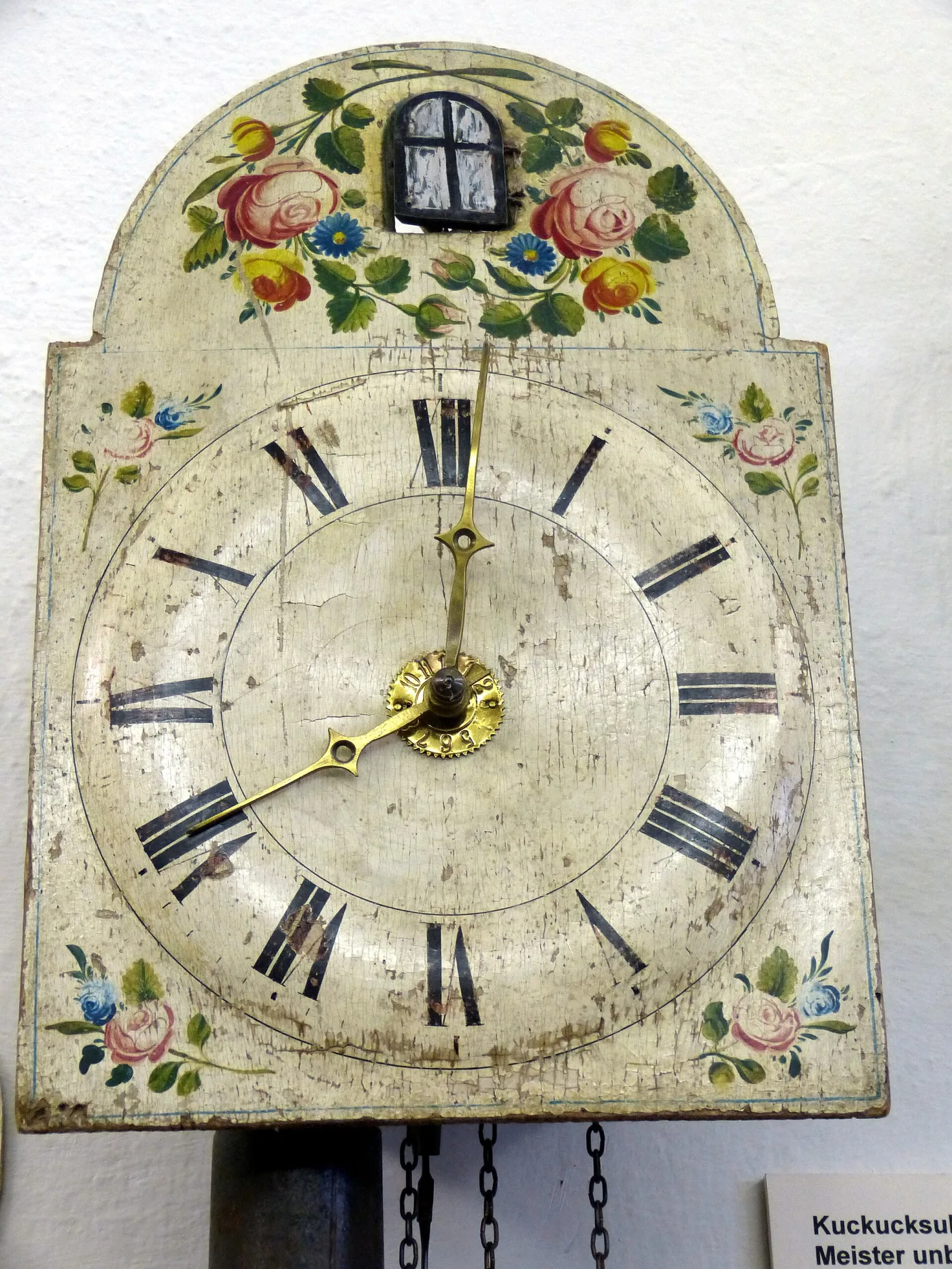 Painted wooden shield clock dial with roses and a small black-framed painted window above the numerals