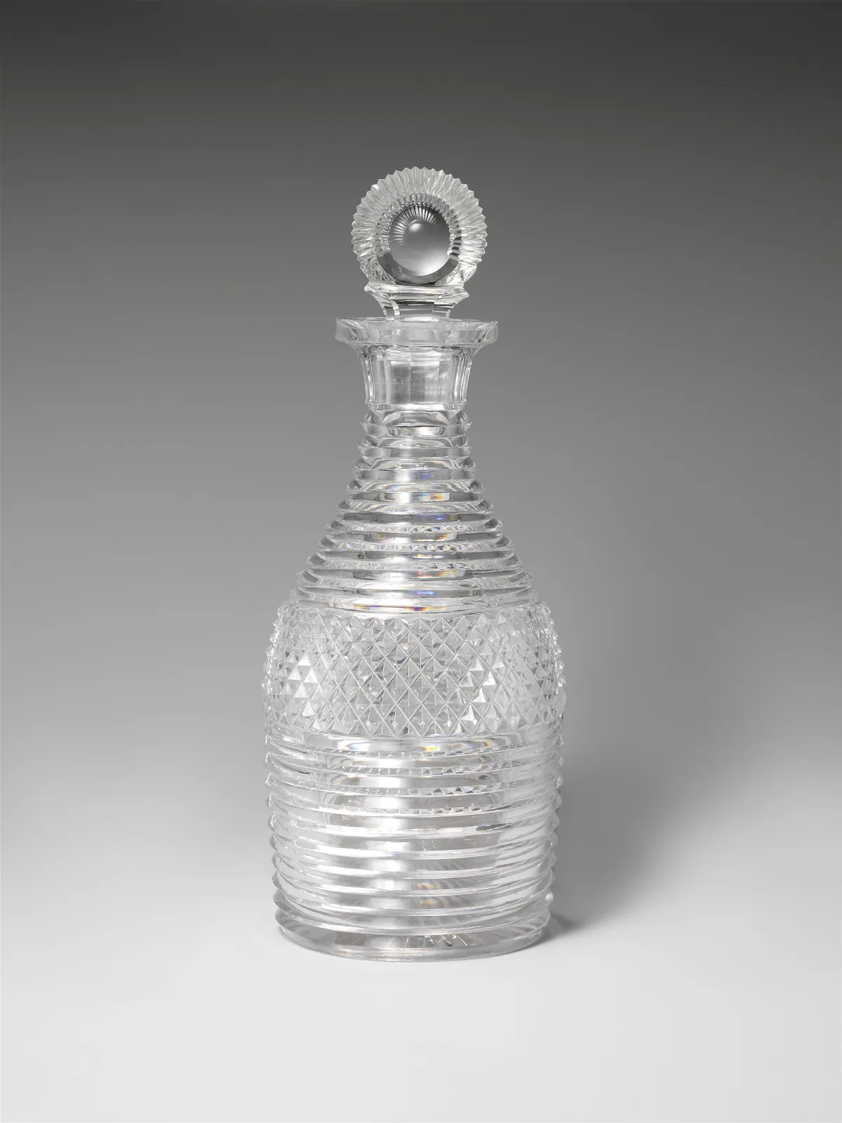 Heavy cut-glass decanter whose entire body is covered in close horizontal step-cut bands, broken at the widest point by a wide band of fine crosscut diamonds, under a fluted flared collar and a flat disc stopper cut in radiating rays around a central bullseye