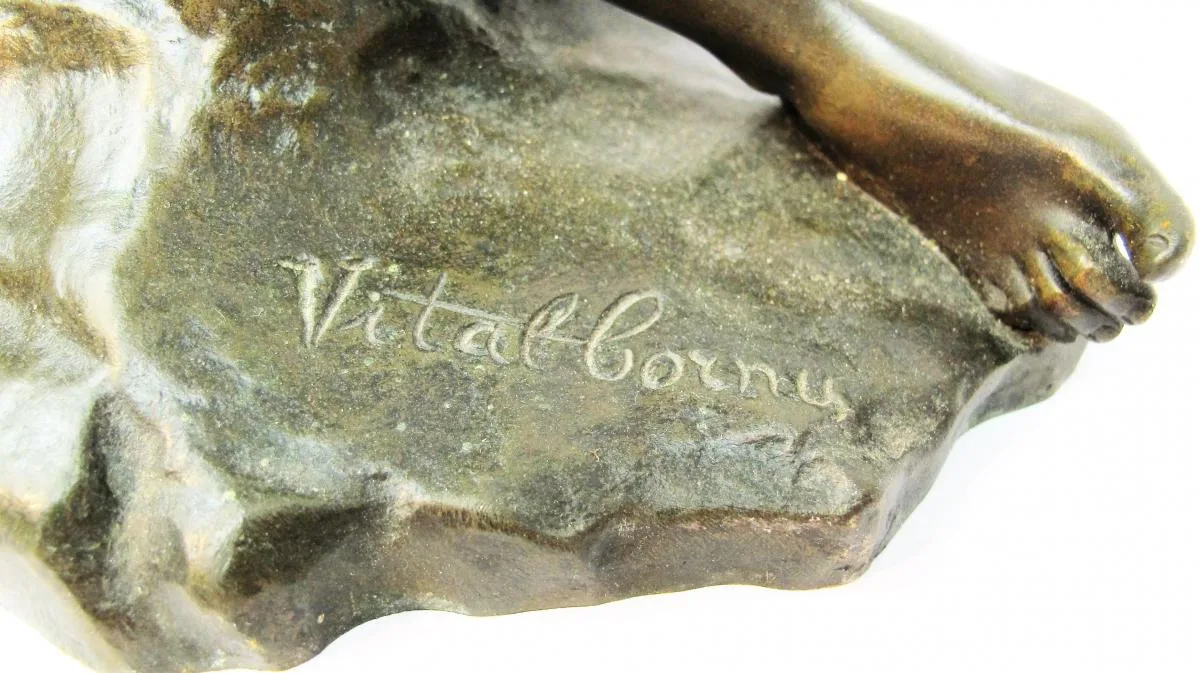 Close view of the rough bronze base of a sculpture beside a bare foot, with a flowing cursive signature reading Vital-Cornu incised into the surface