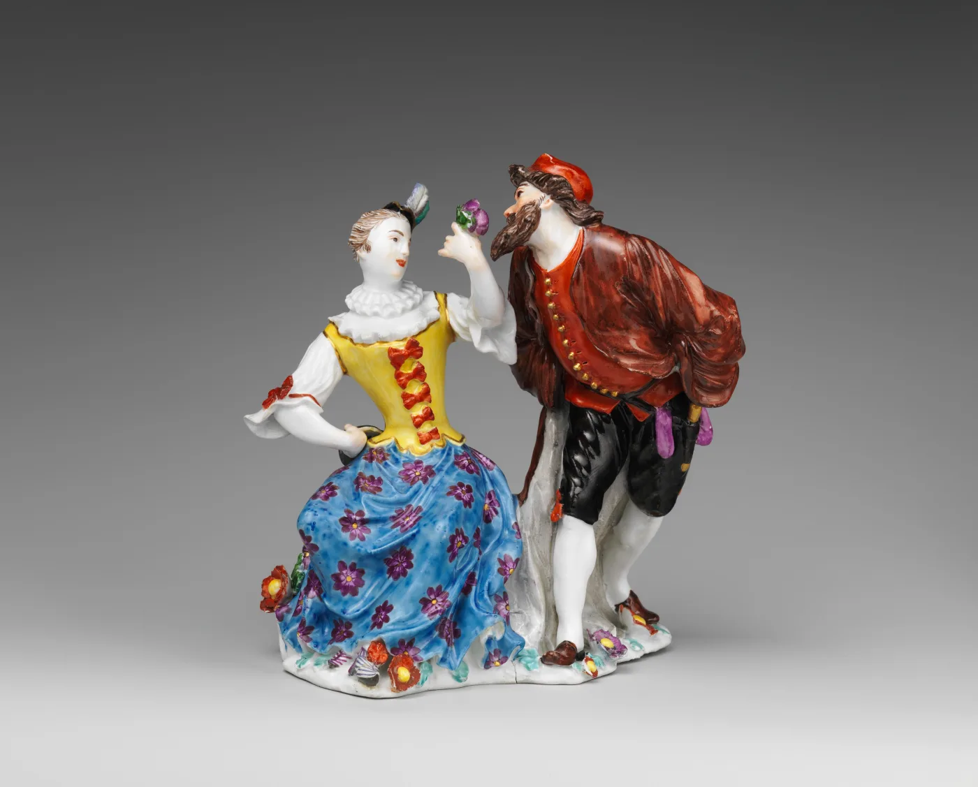 Porcelain group of two figures on a low base: a woman in a striped bodice and flowered skirt turning away, and a bearded man in a red cloak and pointed hat leaning towards her, both brightly enamelled