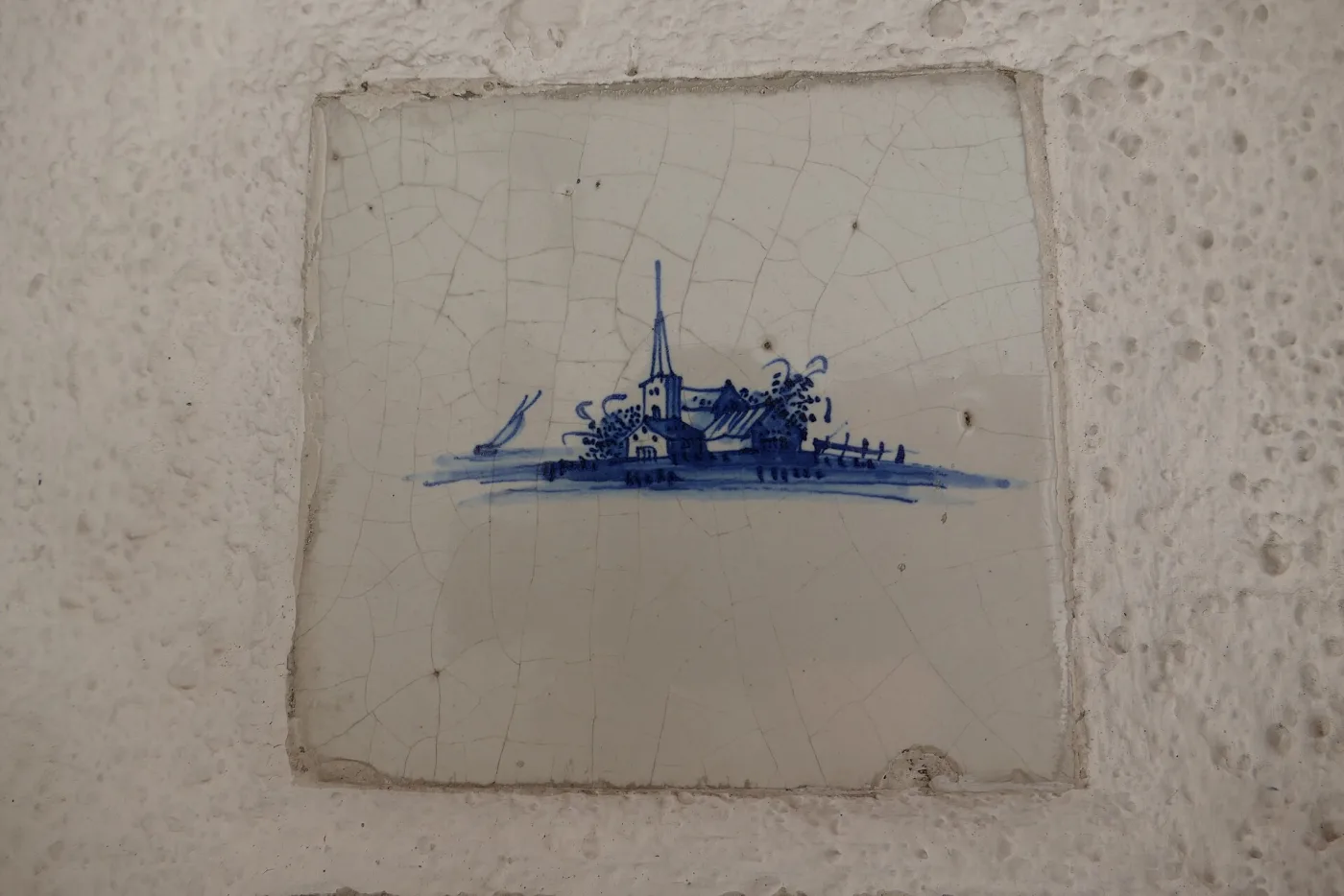 Single blue-and-white Delft tile set in plaster, painted with a small village of houses and a church spire on a low bank, the glaze finely crazed and the tile edges chipped and uneven