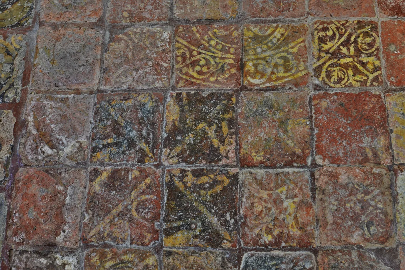 Medieval floor of small square tiles laid in a chequer, alternating dark red and paler bodies, many carrying worn yellow inlaid patterns of foliage and geometric interlace