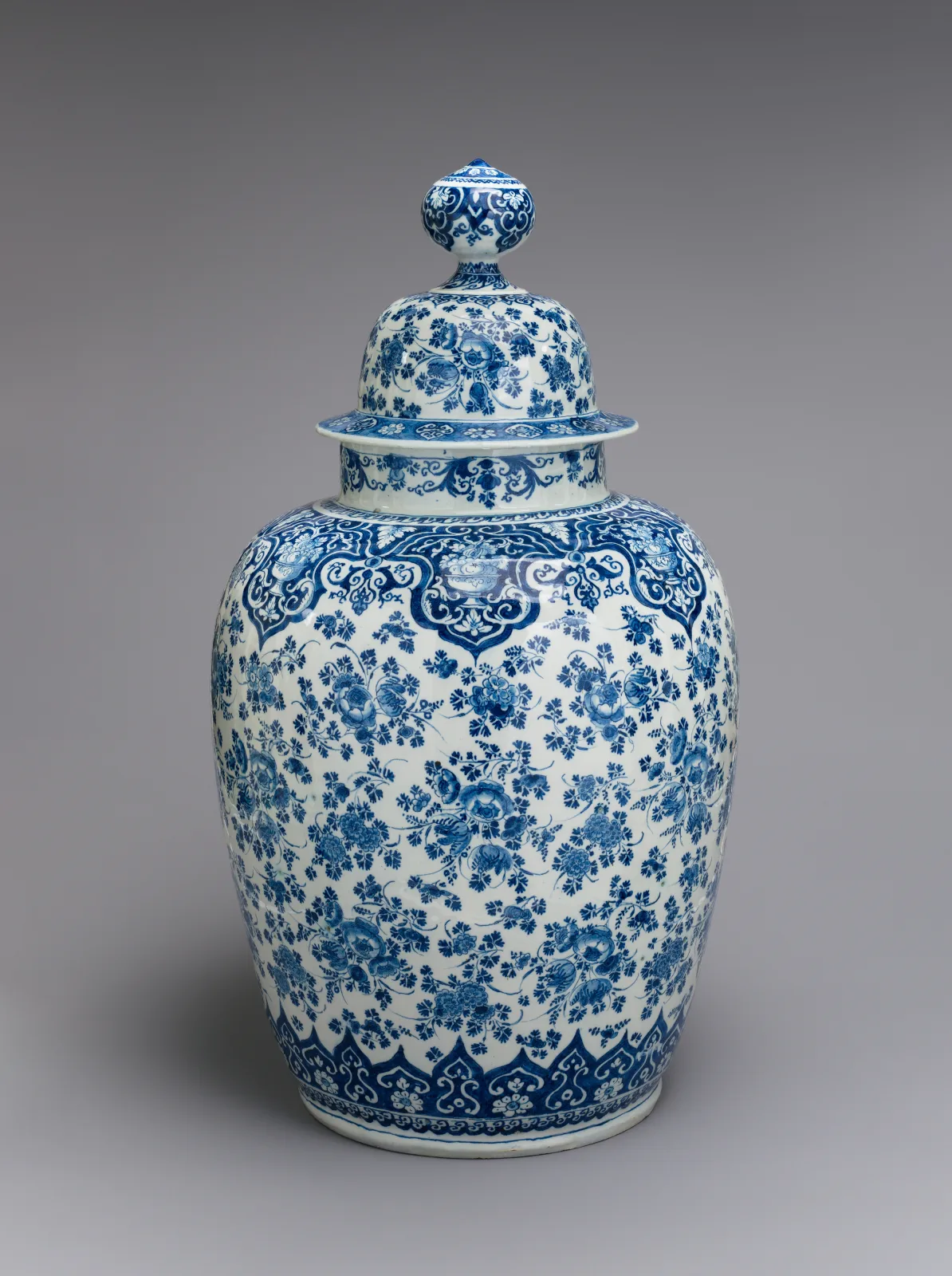 Tall covered baluster vase in white earthenware painted all over in cobalt blue with dense scrolling flowers and foliage, with a band of lappets round the foot and a knopped finial on the domed cover