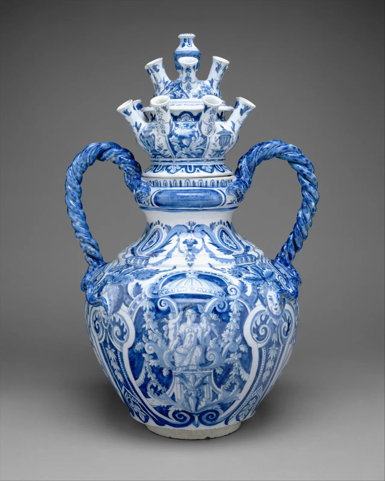 Two-handled blue and white vase with a bulbous moulded body, its shoulder rising into a tier of narrow upturned spouts, the handles formed as twisted ropes and the body panelled with figures under arches