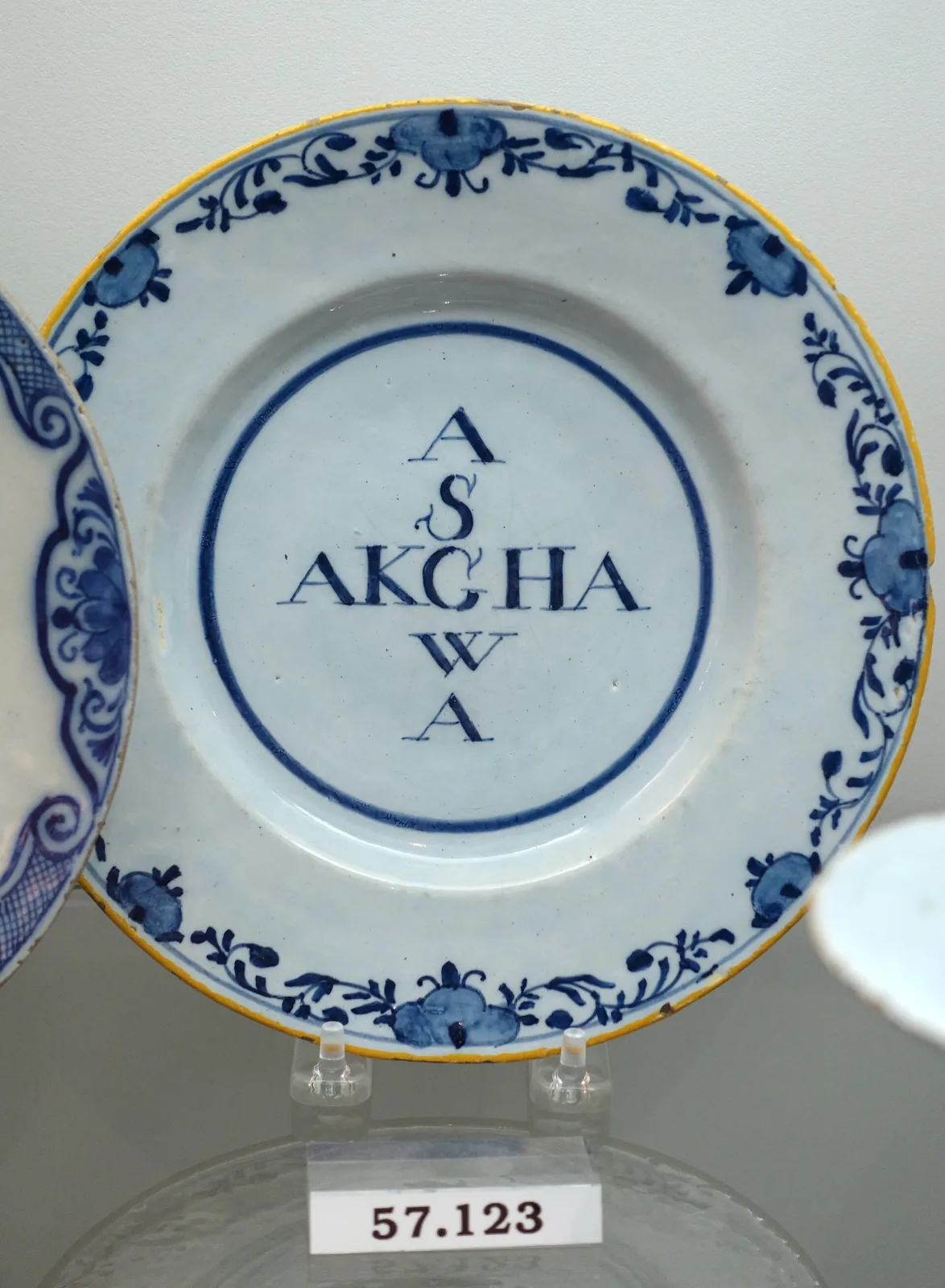 Blue and white plate with a border of flower sprigs and a thin yellow line at the rim, its centre carrying two letter-groups side by side with a single letter stacked above and below each, all inside a plain ring