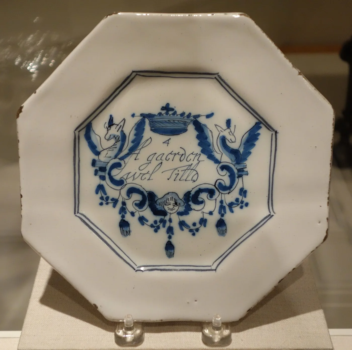Octagonal white plate painted in blue with a cartouche enclosing a line of flowing script, flanked by two birds and hung with swags and tassels below a small crown
