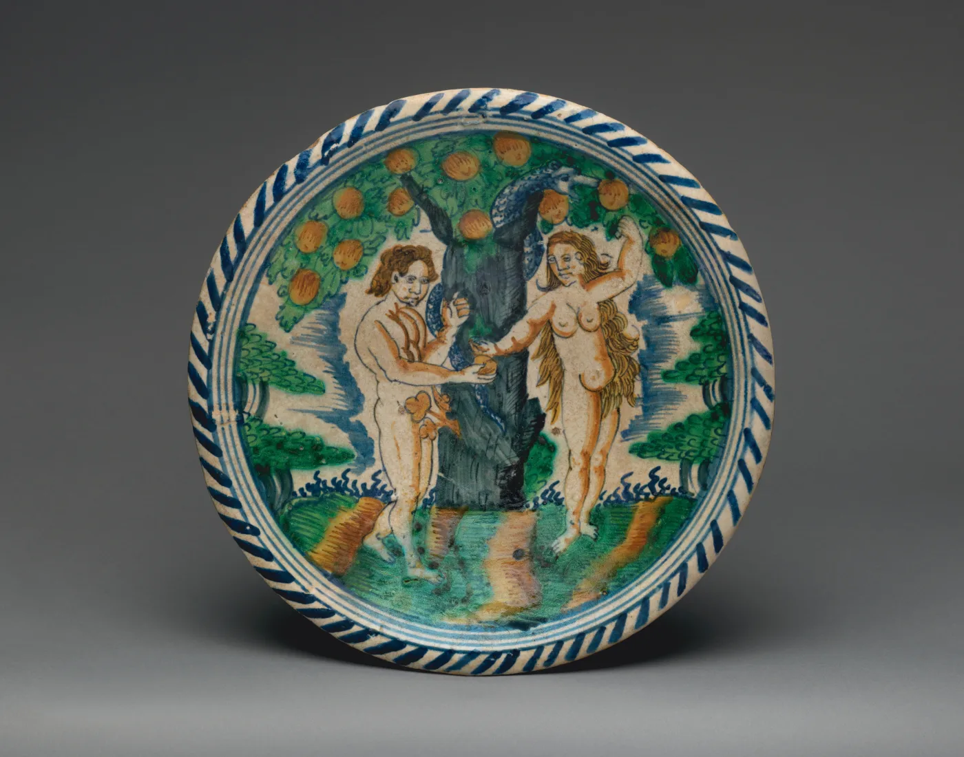 Circular dish painted with Adam and Eve standing either side of a tree hung with orange fruit, in green, orange-brown, yellow and blue, the rim carrying a ring of diagonal blue dashes