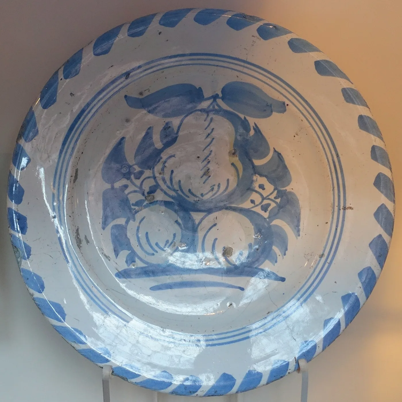 Broad shallow dish painted in pale blue with a loose group of fruit at the centre, its wide rim carrying a ring of short diagonal blue strokes, the glaze chipped in places around the edge