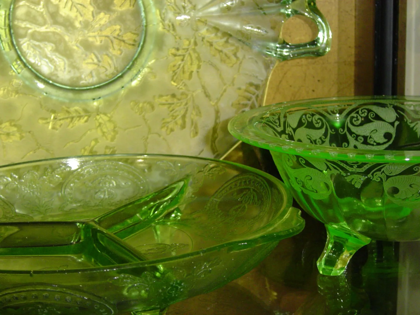 Close view of three pieces of green Depression glass &mdash; a divided relish dish, a footed bowl with a scrolled moulded pattern, and a patterned plate behind them