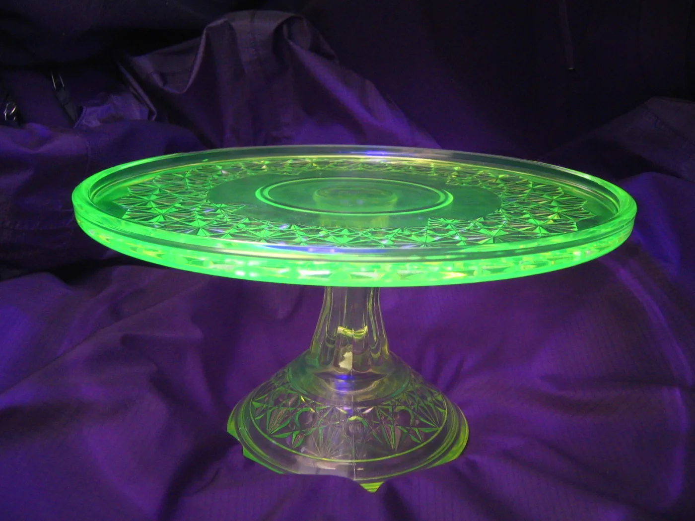 Green glass cake stand on a purple cloth, its pressed pattern glowing bright green under ultraviolet light