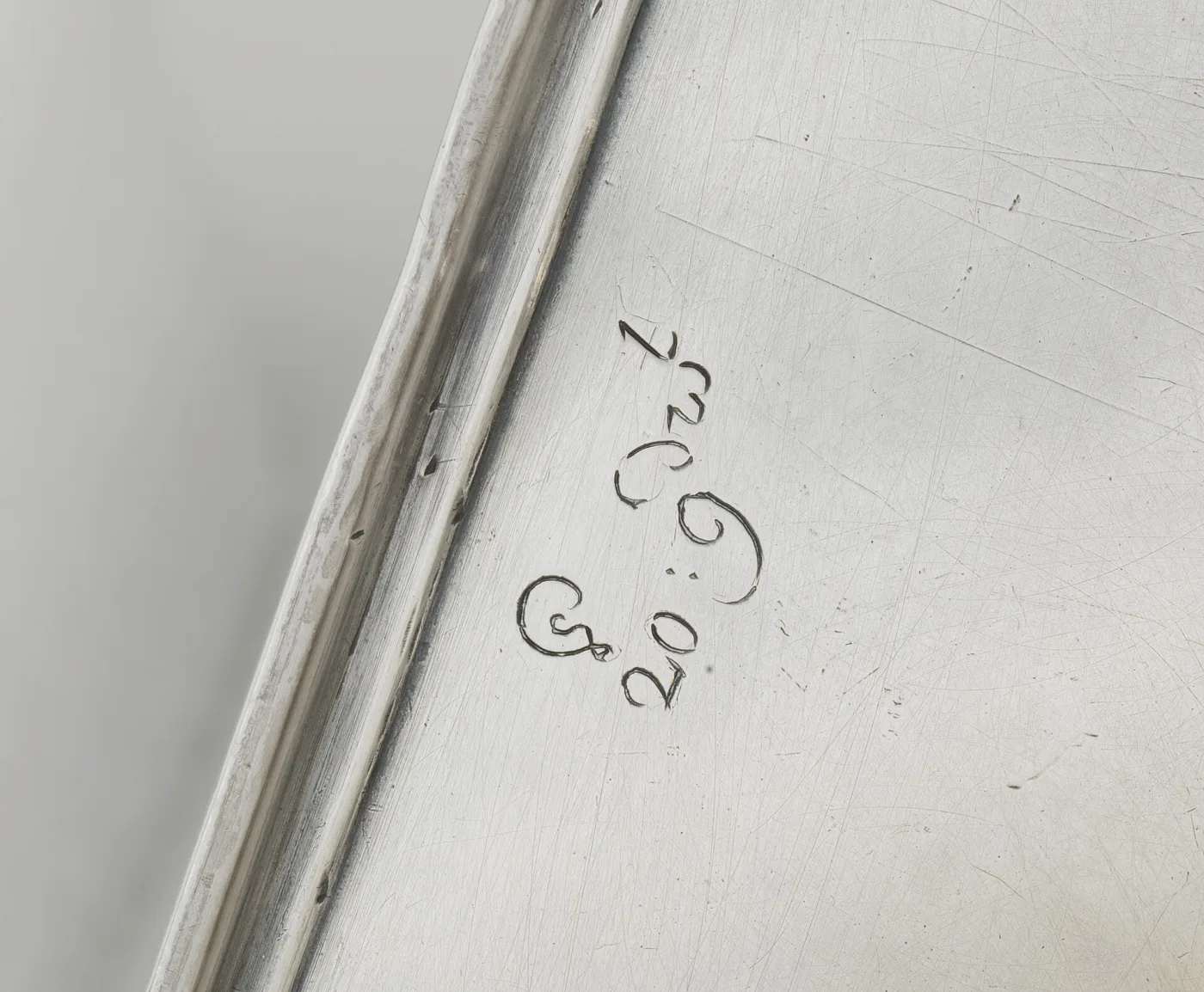 Close view of a scratched silver surface near a moulded edge, engraved with a large flowing cipher of interlaced initials in script