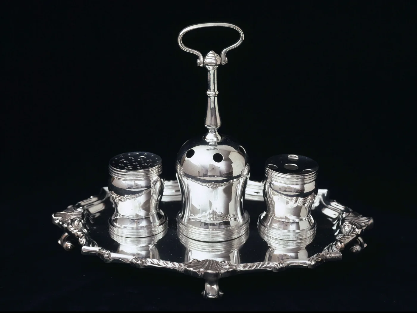 Silver stand on a shaped tray with paw feet and two cast handles, carrying three cylindrical containers — one with a pierced top, one with a single hole, one open — around a tall central shaft with a loop handle