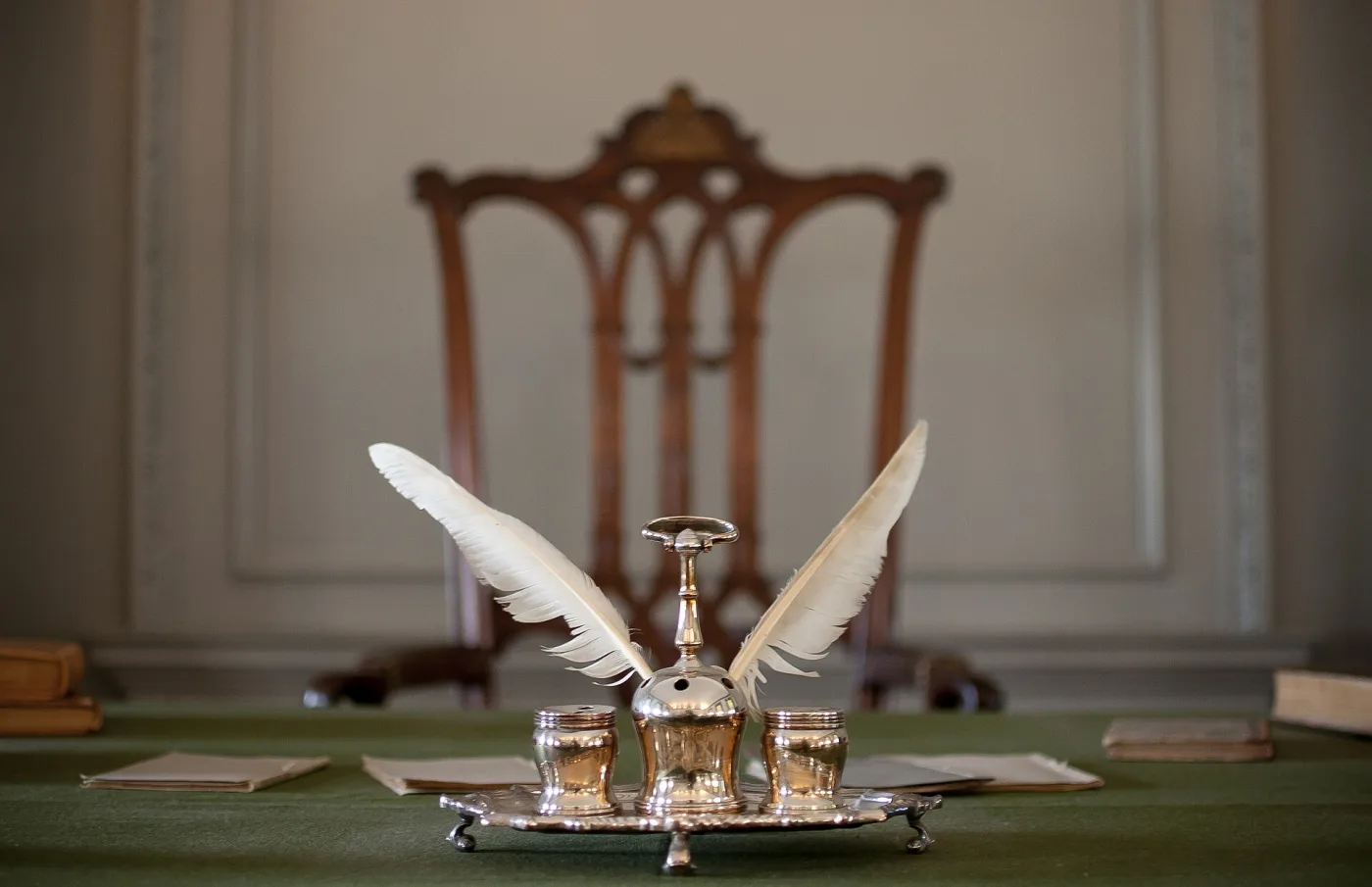 Silver inkstand of the same pattern standing on a green baize table with two large white quills in it, in a panelled room with a carved chair behind