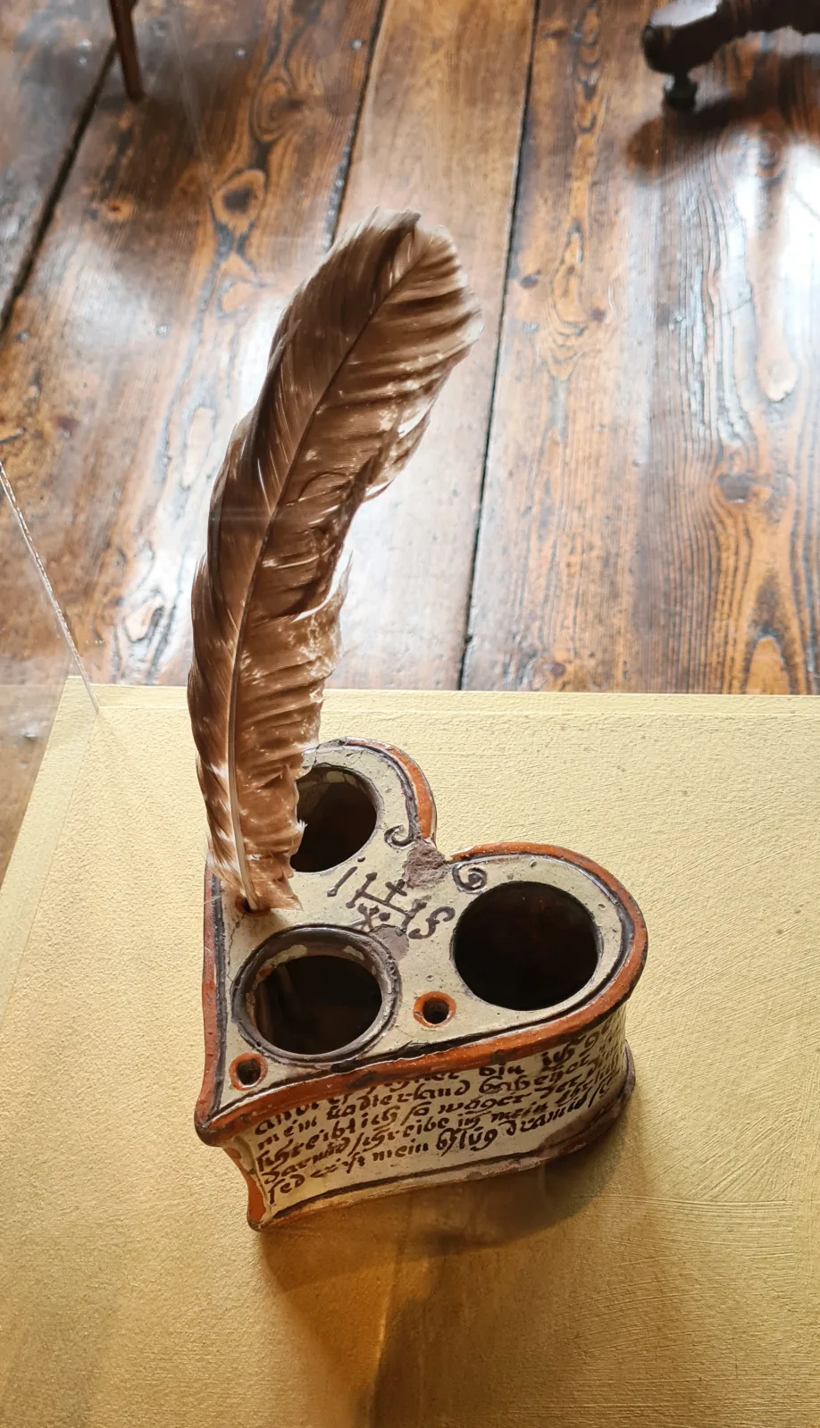 Heart-shaped earthenware stand pierced with three round openings, its sloping front painted with lines of handwritten inscription, a large quill standing in one of the openings