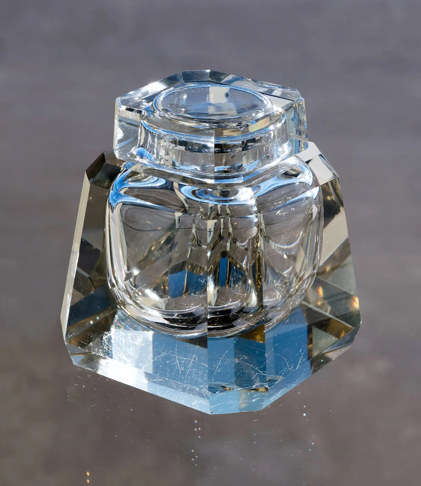 Heavy clear glass inkwell with faceted sloping sides and a hinged metal collar and lid, standing on a plain surface, its surfaces covered in fine scratches