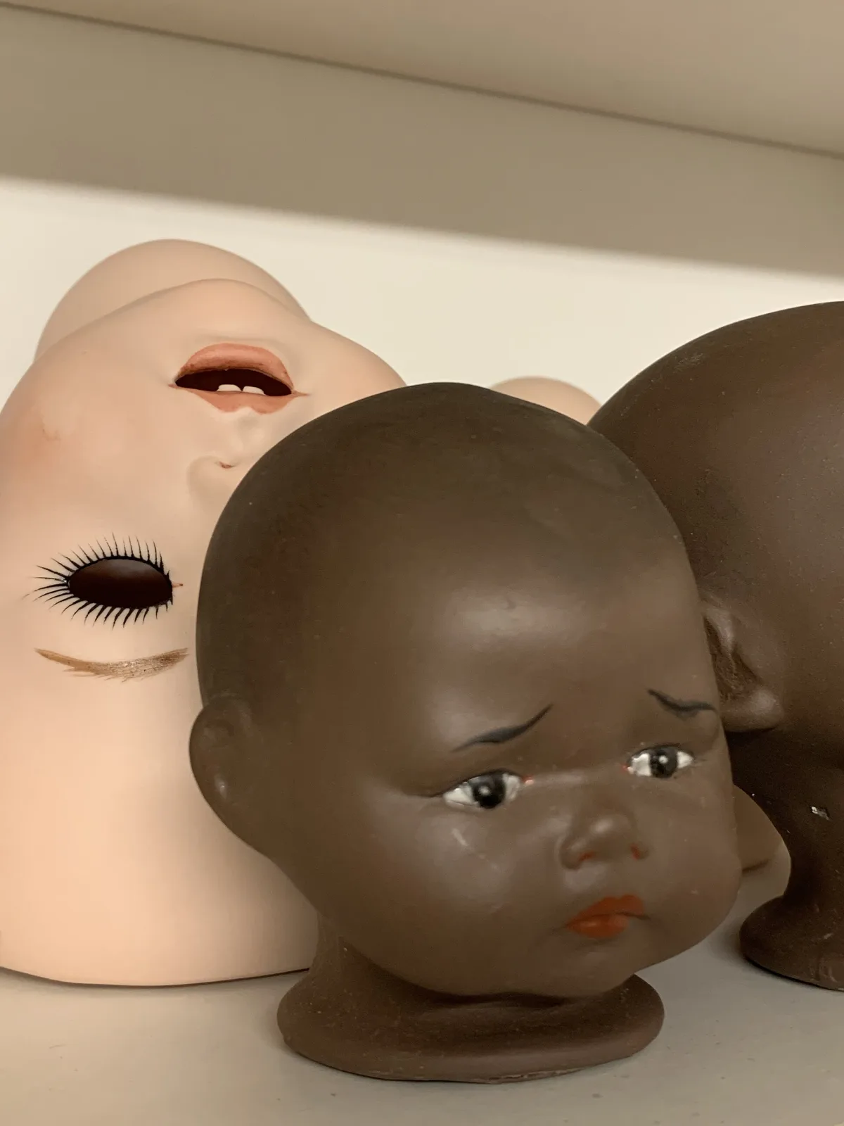 Several doll heads on a shelf: a pale one seen from the side with an open mouth and applied lashes, a dark brown one facing forward with inset eyes and painted brows, and further pale heads behind