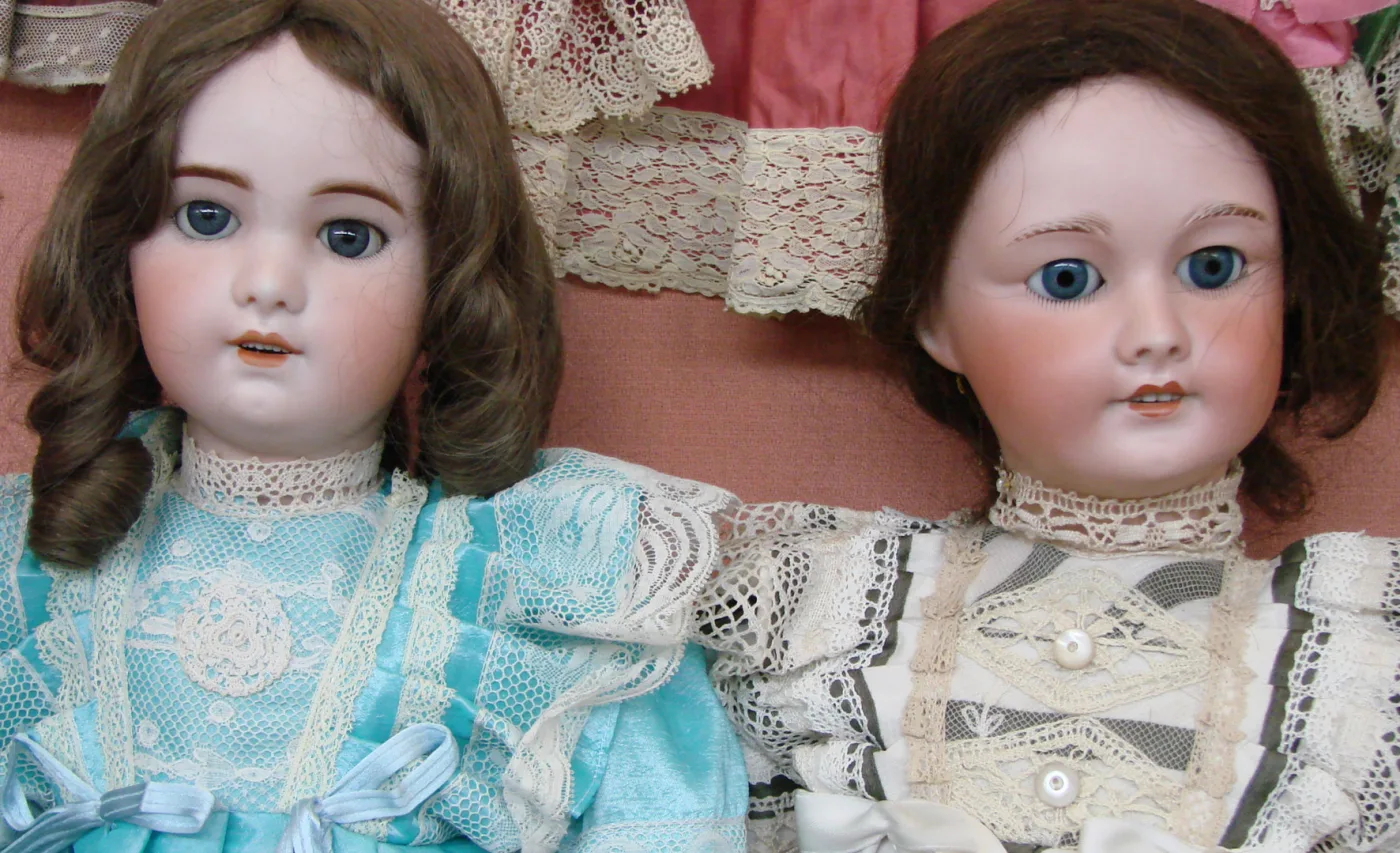 Two bisque-head dolls lying side by side in lace-trimmed dresses, the left with brown eyes and a slightly open mouth showing teeth, the right with bright blue eyes and a closed mouth