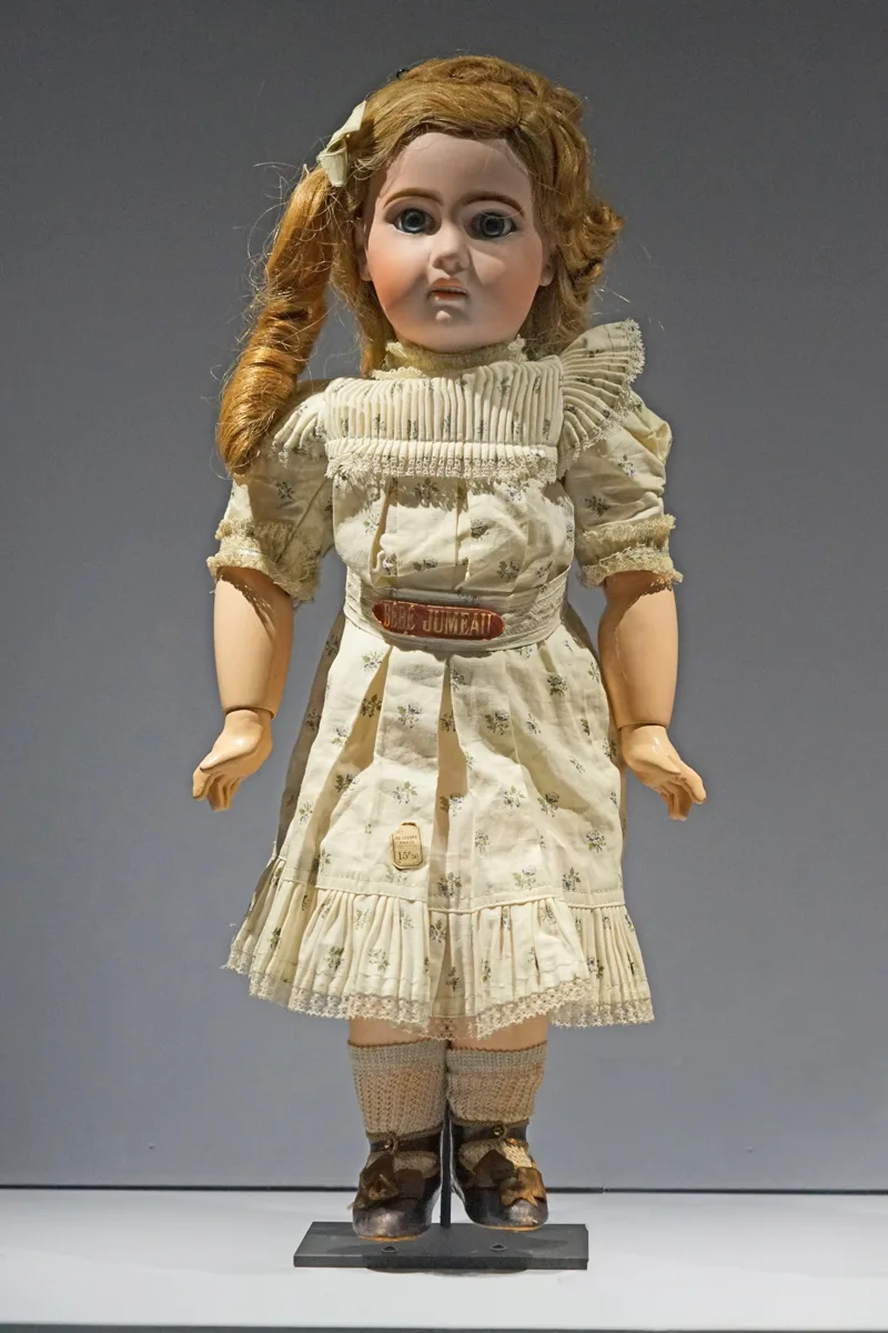 Bisque-head girl doll standing on a museum mount in a cream sprigged dress, with a long fair wig, large grey glass eyes, ball joints visible at the elbows of its composition arms, and a red and gilt belt plaque lettered Bébé Jumeau at the waist