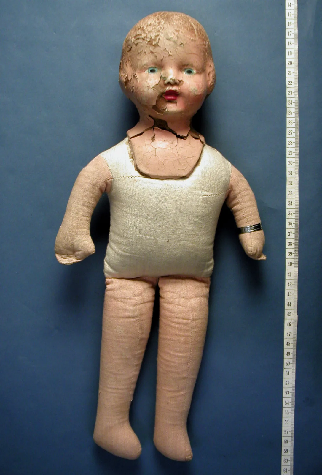 Doll with a moulded composition head and lower limbs on a plain stuffed cloth torso, laid flat beside a vertical ruler that shows its full height