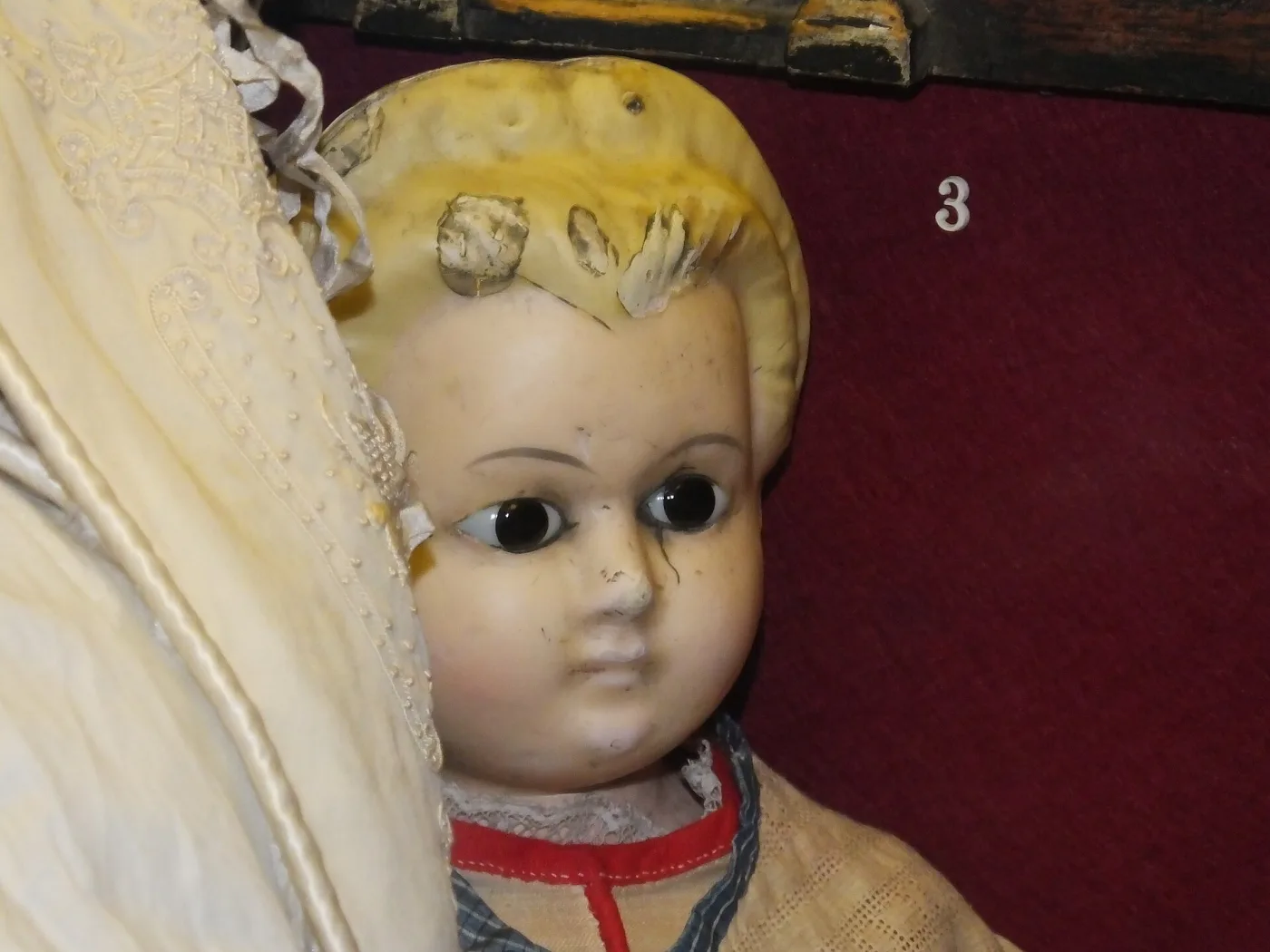 Close view of a doll's head of wax over a substrate, its yellow moulded hair and pale face broken by wide flaking cracks that lift away from the surface, with dark inset glass eyes, lying in a lined display case