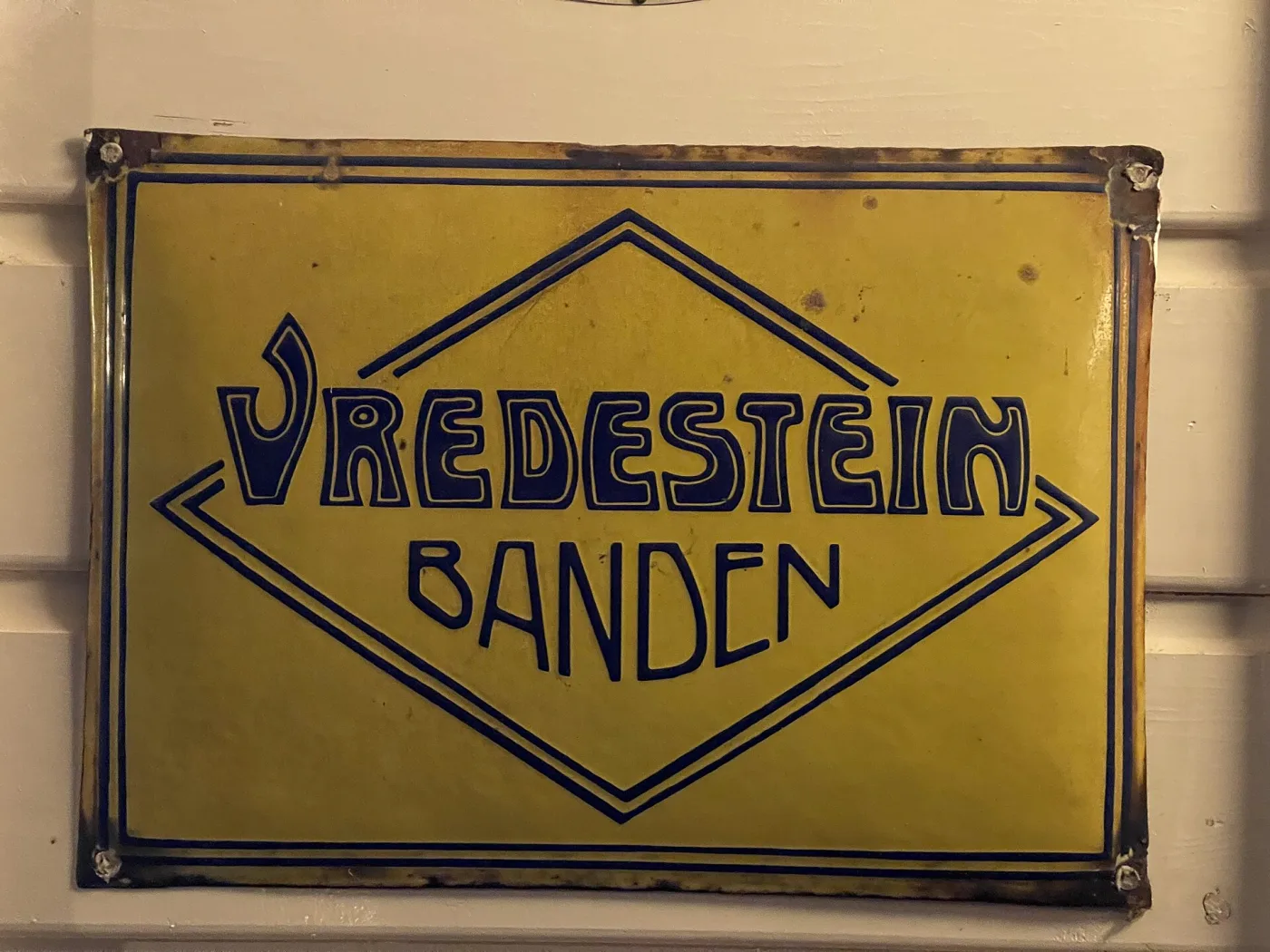Diamond-shaped enamel sign on a yellow ground lettered Vredestein Banden in dark blue, its four corners pierced with mounting holes and its edges chipped and rust-stained