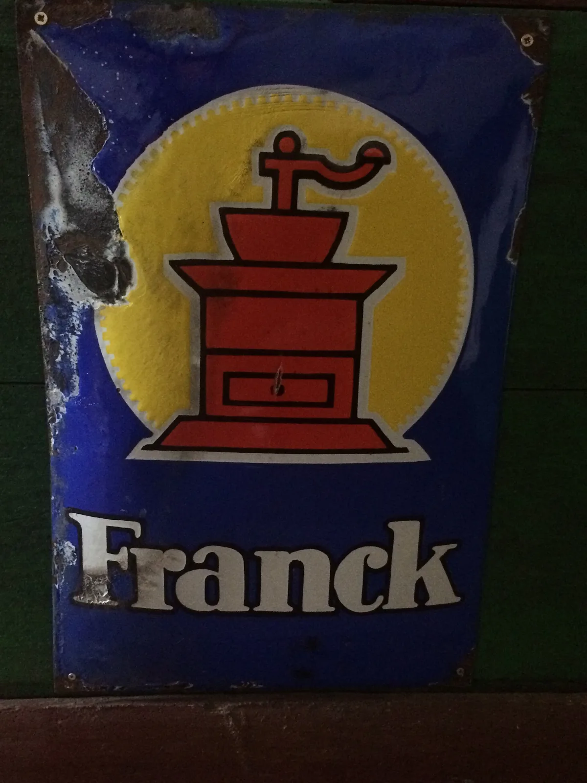 Enamel sign on a deep blue ground showing a red coffee grinder against a yellow disc, its top corners and upper edge broken away in large irregular patches to expose dark bare steel beneath the enamel