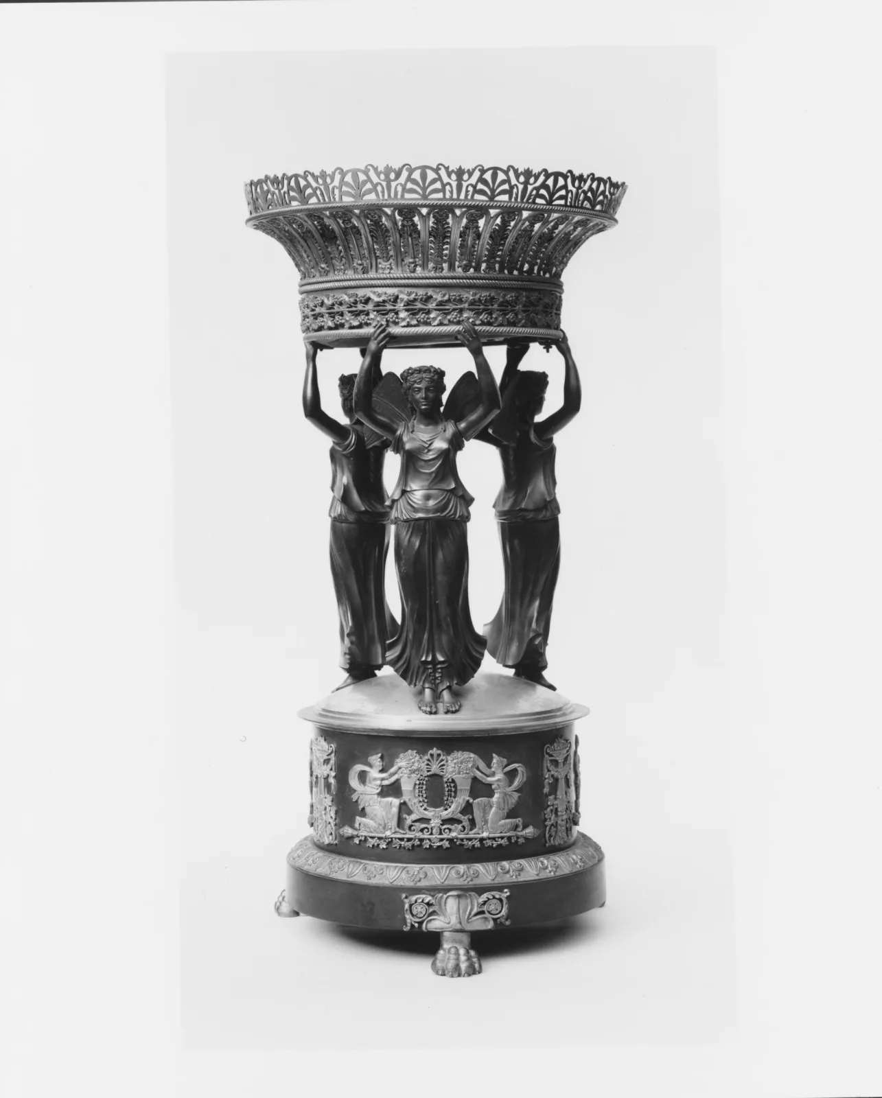 Tall silver-gilt epergne whose deep pierced bowl is supported by three standing classical figures on a drum-shaped plinth decorated with a frieze, the whole raised on paw feet