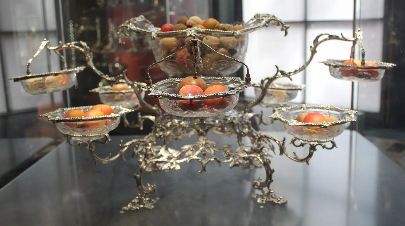 Silver epergne in a museum case with a central basket and four hanging baskets on scrolled branches, filled with fruit and glass dishes as it would have been used at table