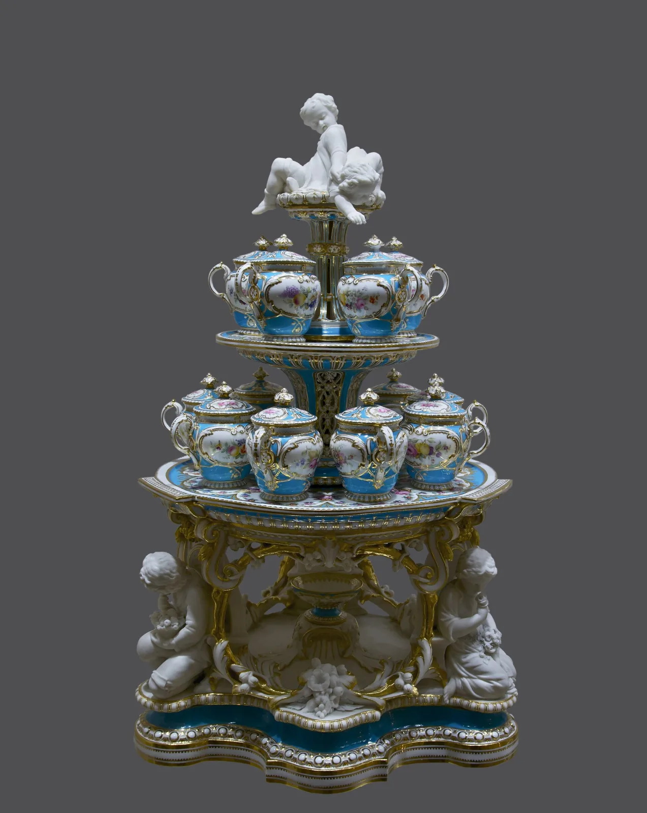Tall tiered dessert stand of blue and white porcelain and gilt bronze, its three graduated levels carrying matching covered cups and dishes, topped by a small white figure