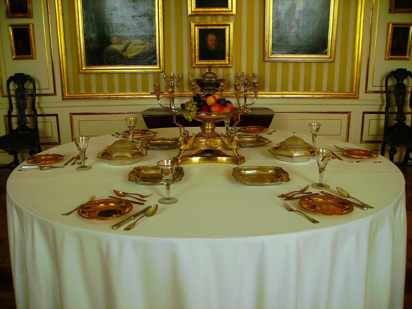 Formal dining room hung with gilt-framed pictures, its round table laid with white cloth, gold-mounted place settings and covered dishes, with a gilt epergne of fruit at the centre