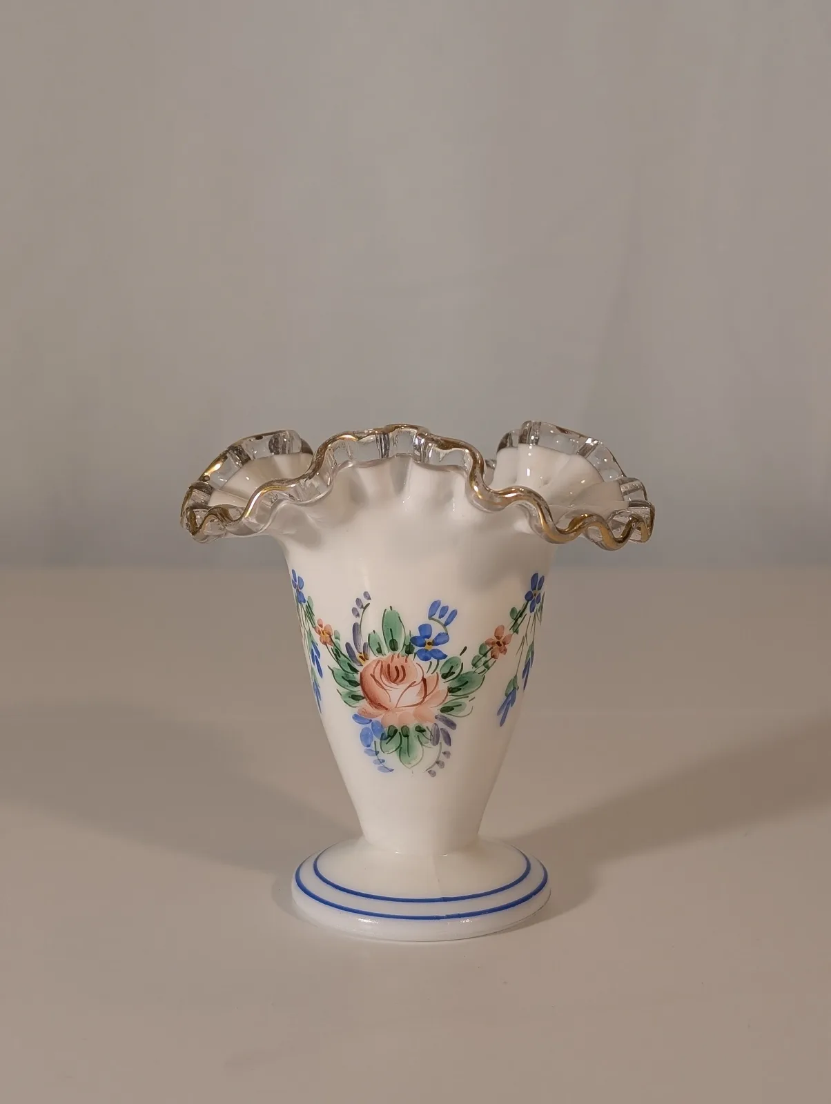 Small milk-glass vase with a clear crimped ruffle, hand-painted with a spray of pink roses and blue flowers, standing on a foot banded in blue