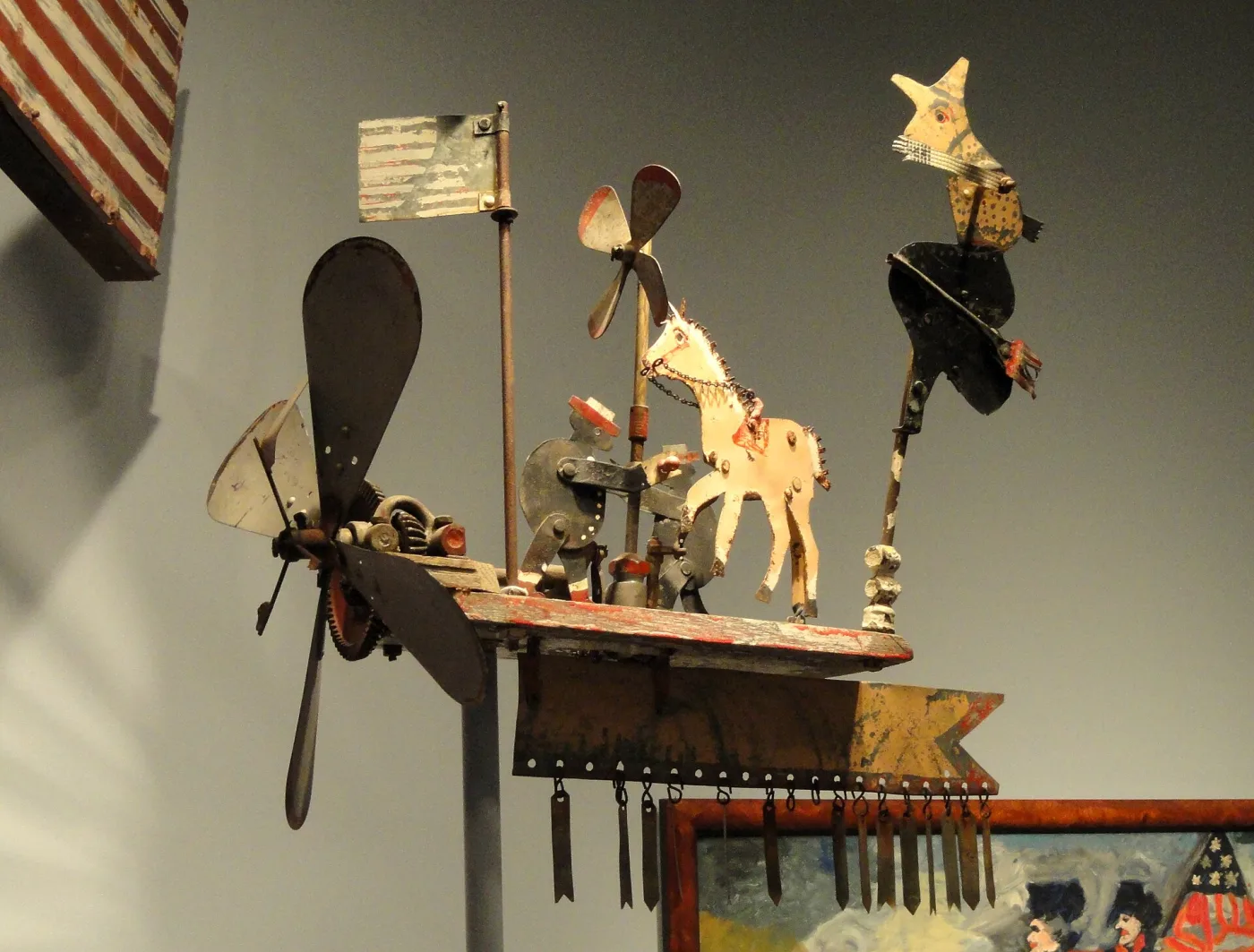 Whirligig with two propellers, a sheet-metal flag, a white horse, a standing figure and a fringe of hanging metal tags on a painted plank platform