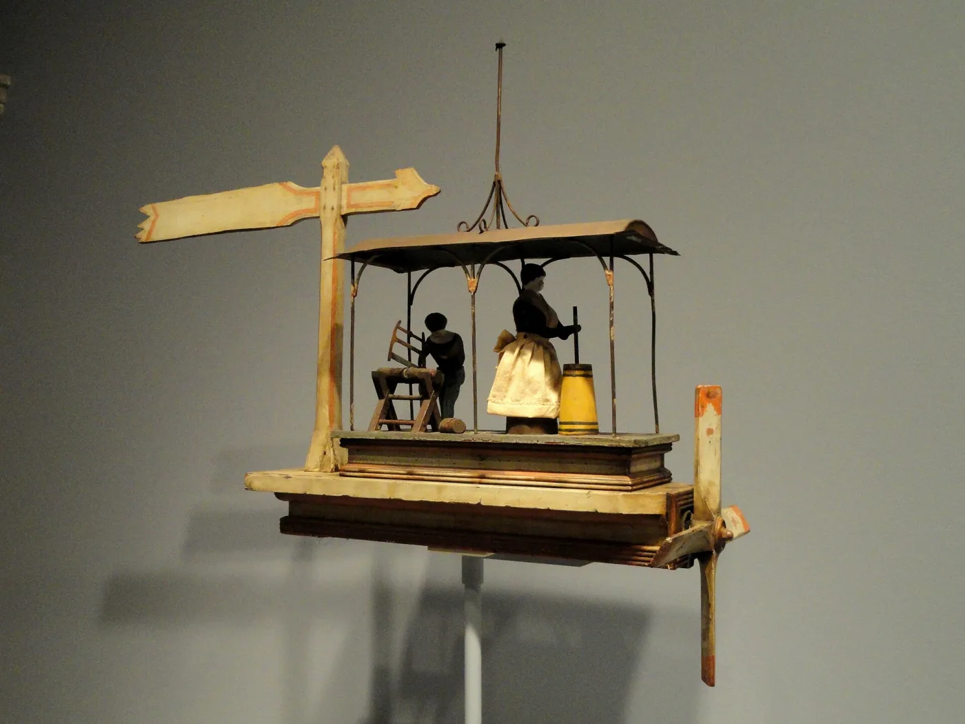 Whirligig modelled as a roofed shed in which a woman works a butter churn and a man saws wood, with a signpost arm and a small propeller