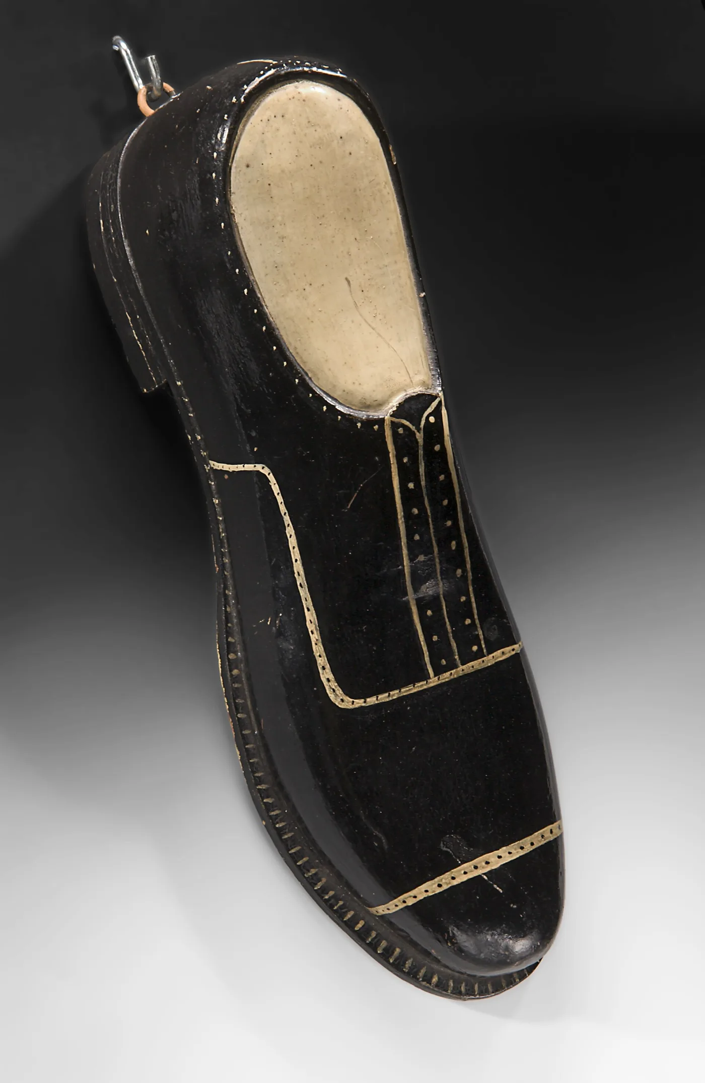 Black-painted wooden shoe used as a shoemaker's sign, with cream lines and dots imitating stitching and welt, and an iron hanging ring at the heel
