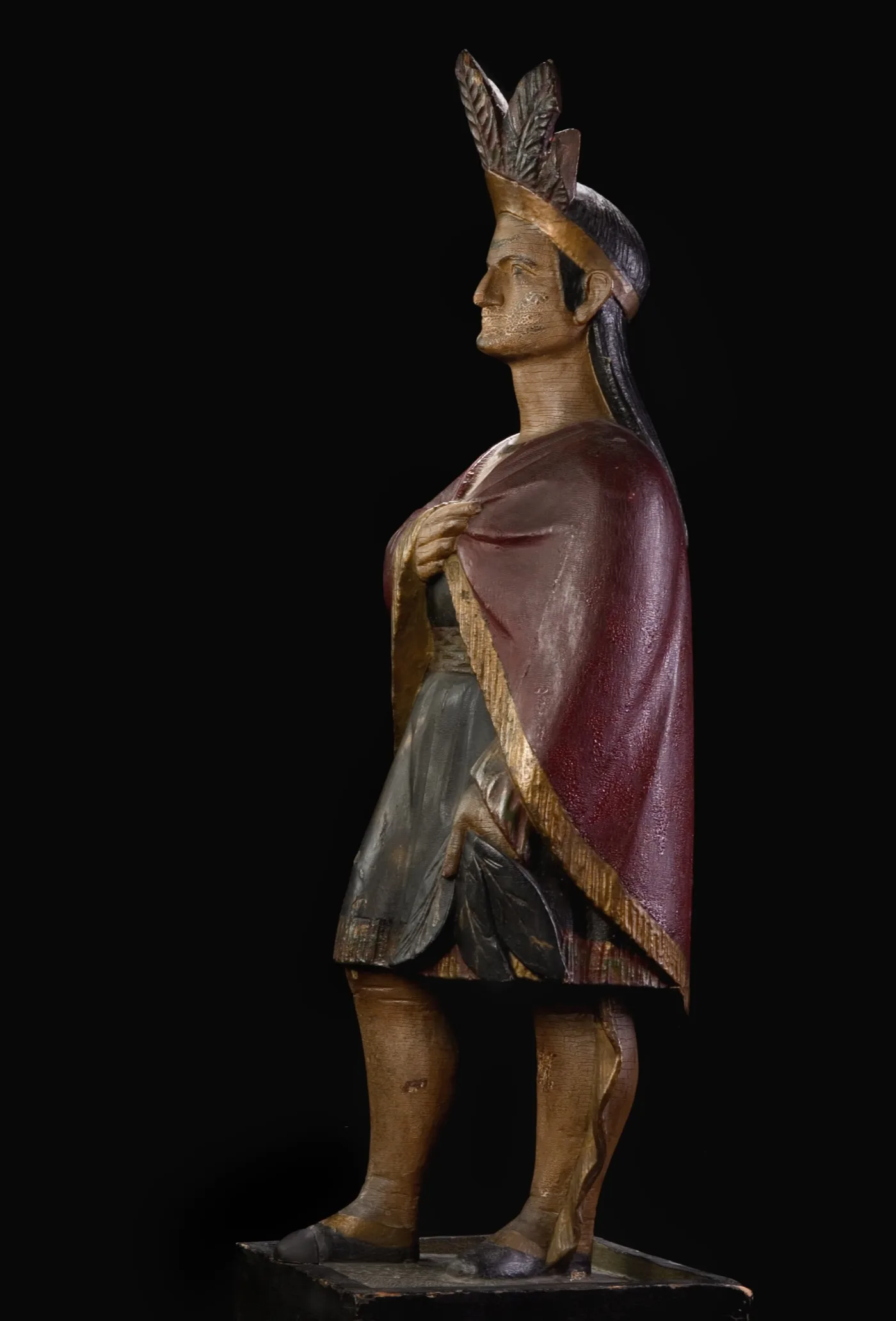 Carved and painted wooden tobacconist's figure of a woman in a feathered headdress and a red cloak, shown in profile on a plinth